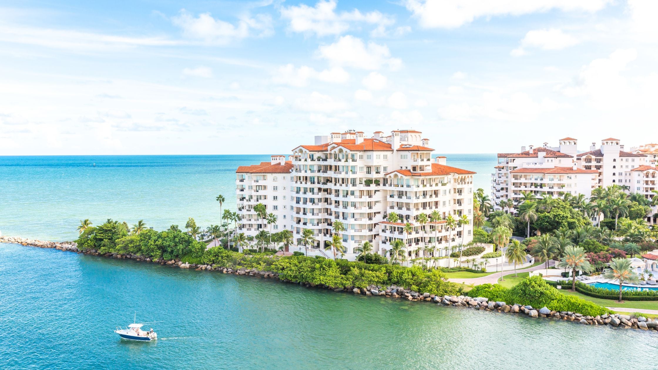 Aerial view of luxury Miami waterfront condo buildings with red tile roofs, palm trees, and turquoise bay water.