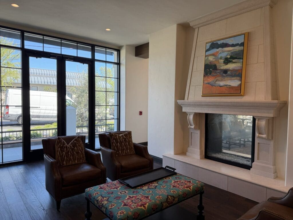 Sitting area at Aegis Living San Rafael with two leather armchairs, an upholstered ottoman and a stone-mantel see-through fireplace beneath a landscape painting