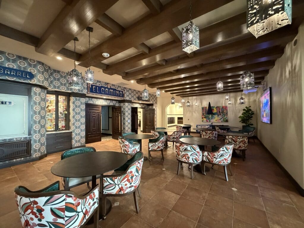Cafe and bistro seating area at Aegis Living San Rafael with floral chairs, dark wood ceiling beams, pierced metal pendant lights and an El Camino storefront sign