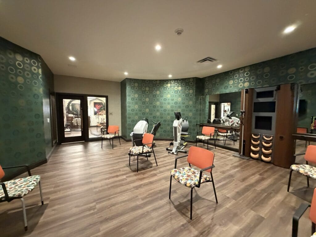 Exercise room at Aegis Living San Rafael with teal patterned wallpaper, orange chairs, recumbent cross-trainers and a mirrored wall