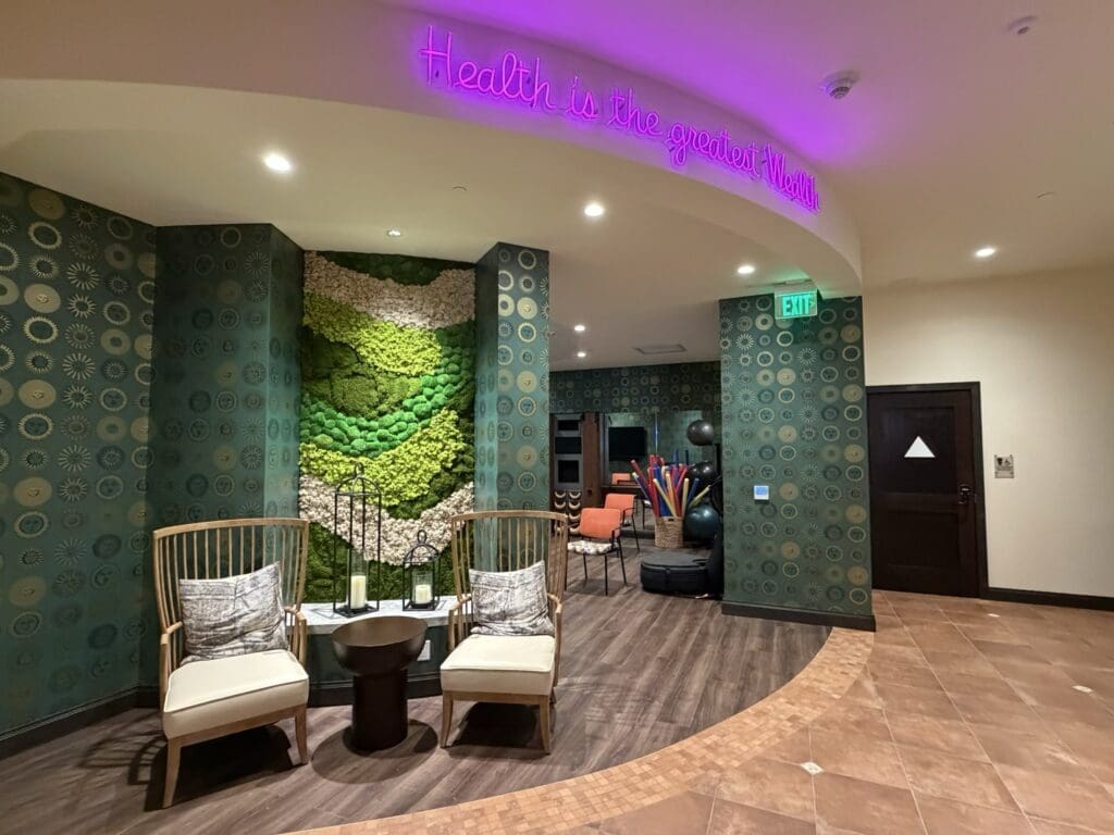 Wellness area entrance at Aegis Living San Rafael with a pink neon sign reading Health is the greatest wealth, a green moss wall and two lounge chairs