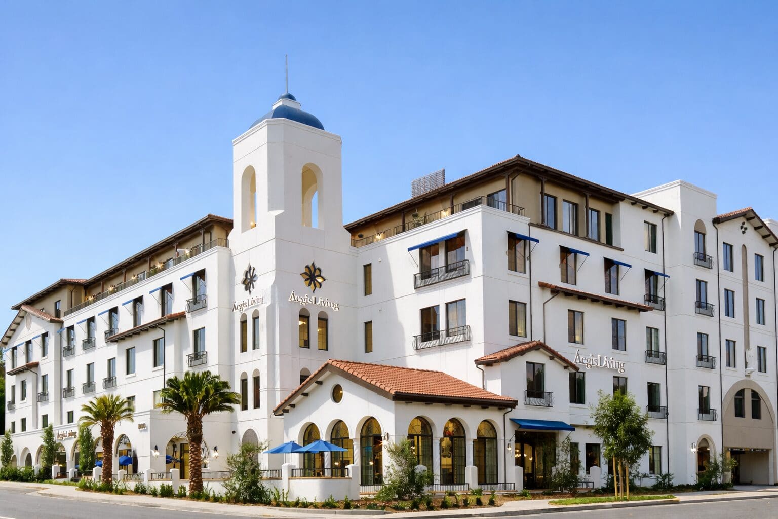 Exterior of Aegis Living San Rafael, a white Mission Revival style building with a blue-domed bell tower, red tile roofs, arched windows and palm trees on Mission Avenue