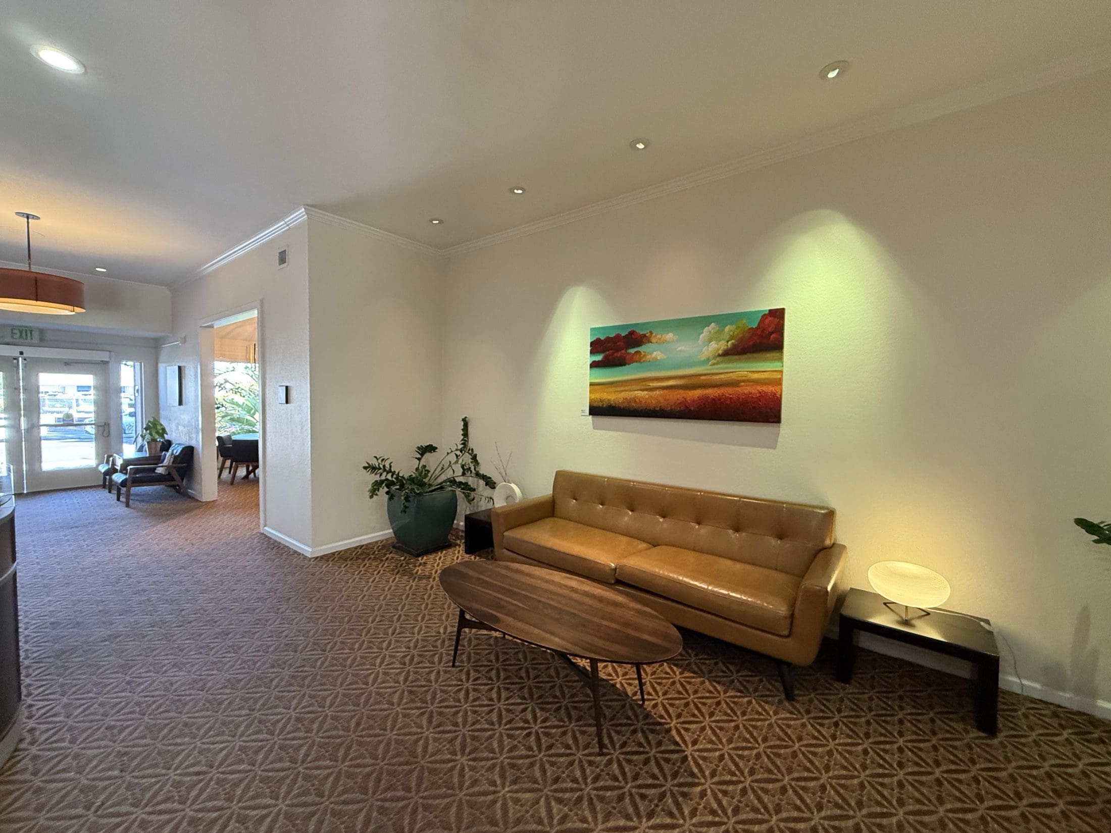 Atria Willow Glen entry lounge with a tan leather sofa, oval wood coffee table, potted plants and a large landscape painting, with glass front doors in the background