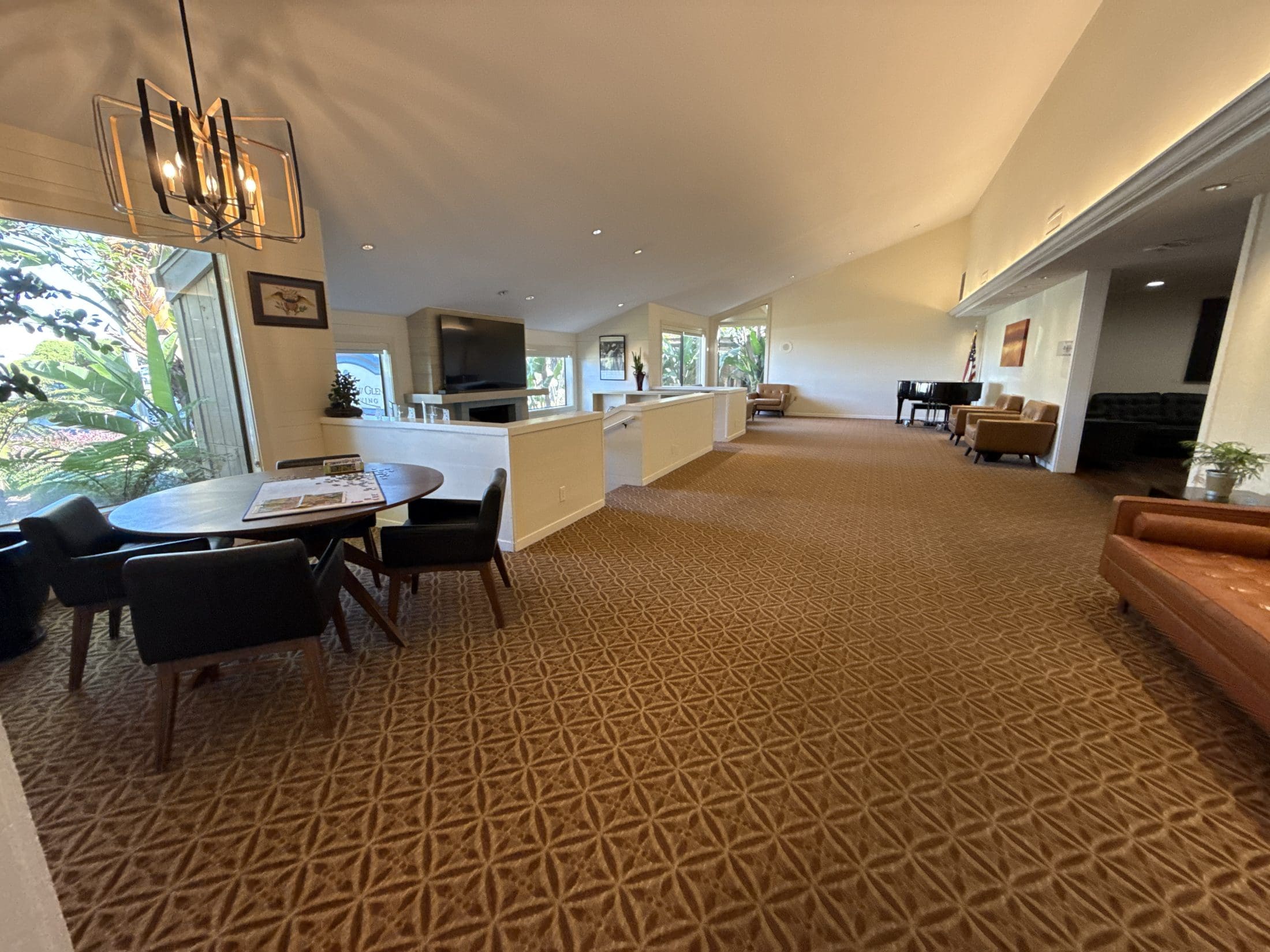 Great room at Atria Willow Glen with a vaulted ceiling, modern chandelier, fireplace with television, grand piano, leather armchairs and a round table with a jigsaw puzzle