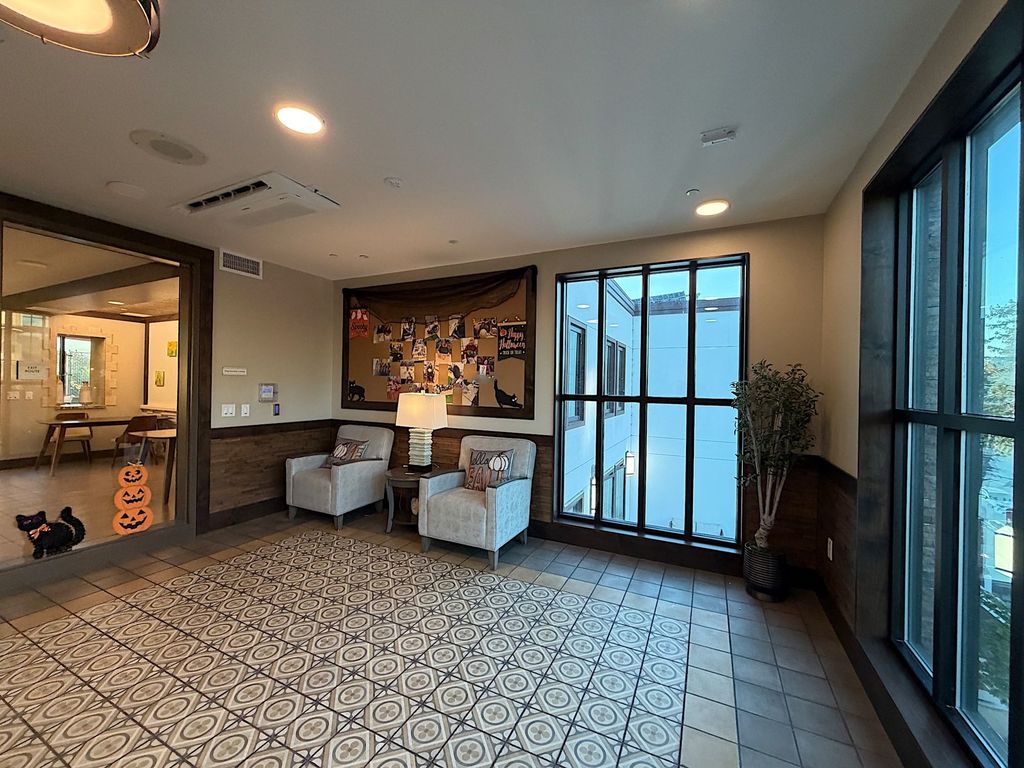 Lounge with armchairs, floor-to-ceiling windows and seasonal resident photo board at Villa Toscana Memory Care