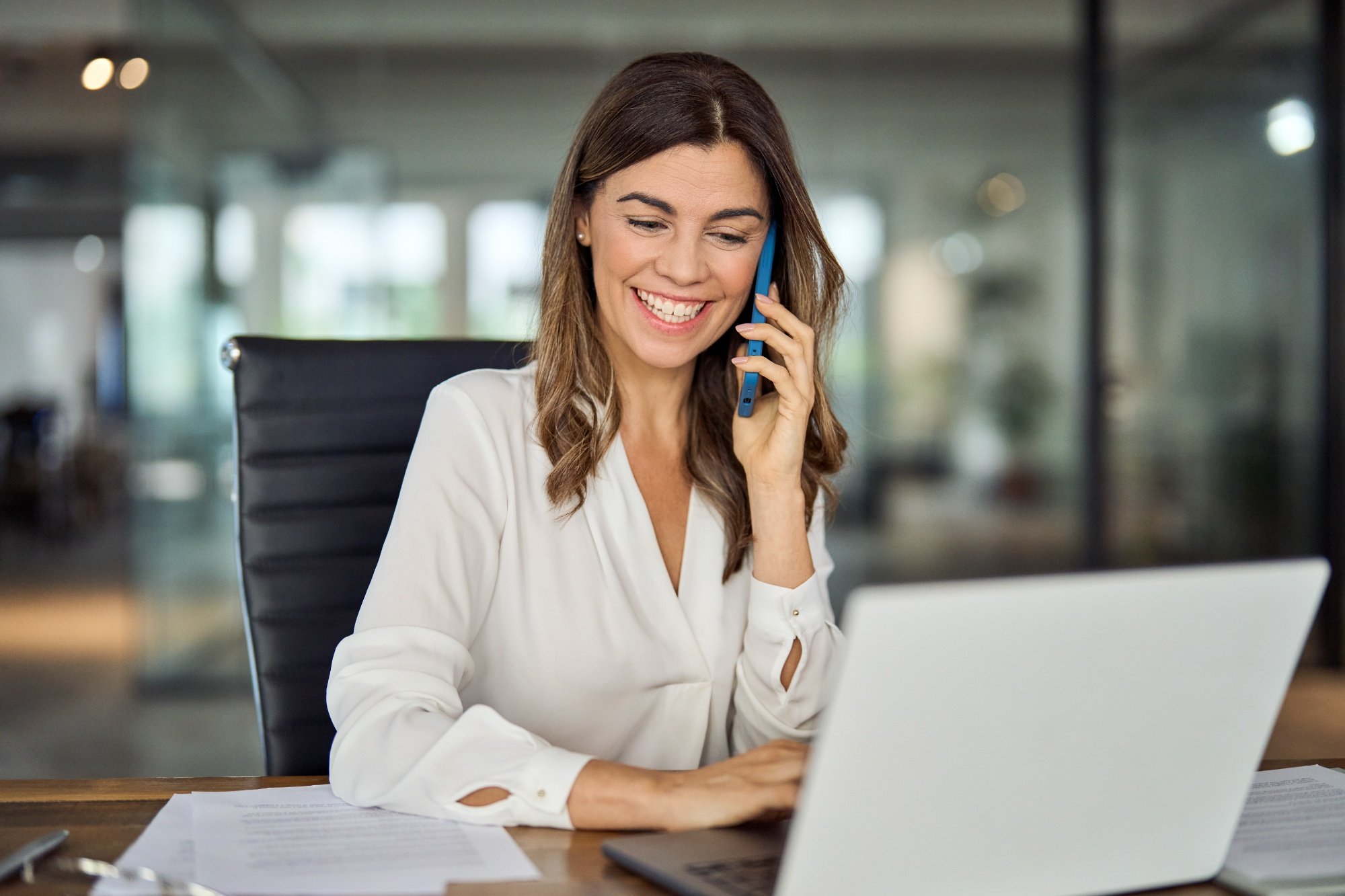 Happy smiling mature middle aged business woman, 40s professional lady executive manager talking on the phone making business call on cellphone at work in office using laptop computer. (1)
