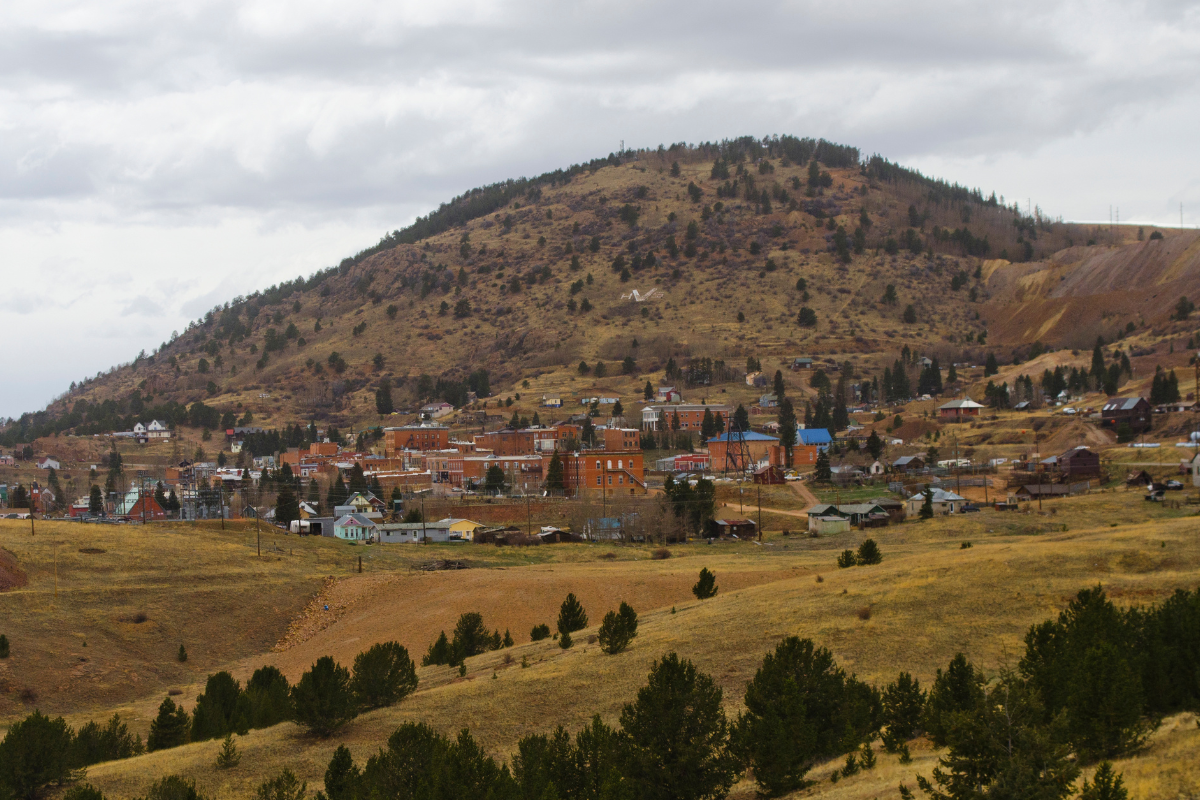 A scenic view of Victor, Colorado, a historic gold mining town surrounded by rolling hills and pine trees. The photo shows old brick buildings, mining structures, and mountain terrain under a cloudy sky.