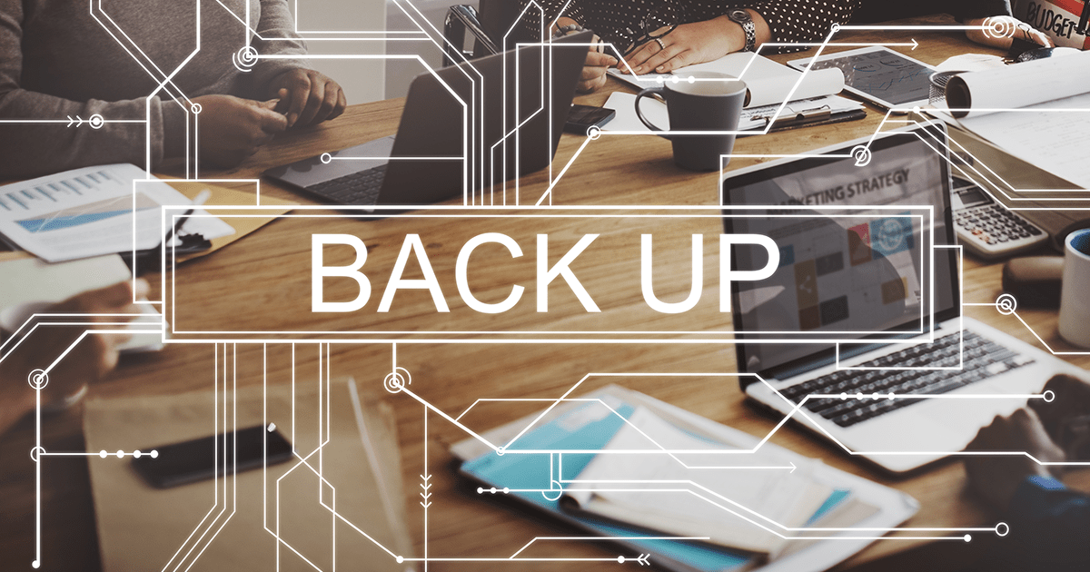 Image of wooden table top with paperwork and laptops. Copy over image says "Back Up"