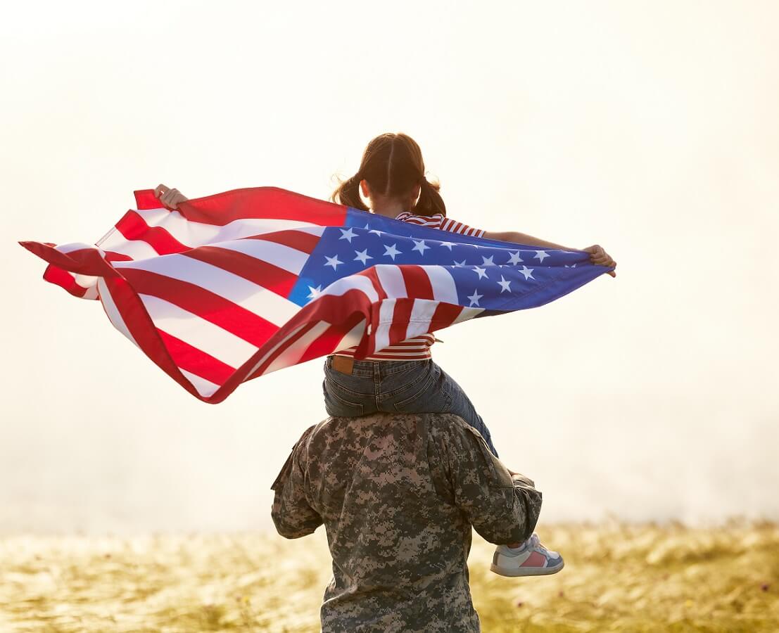 Rear view of military man father carrying happy little daughter with american flag on shoulders
