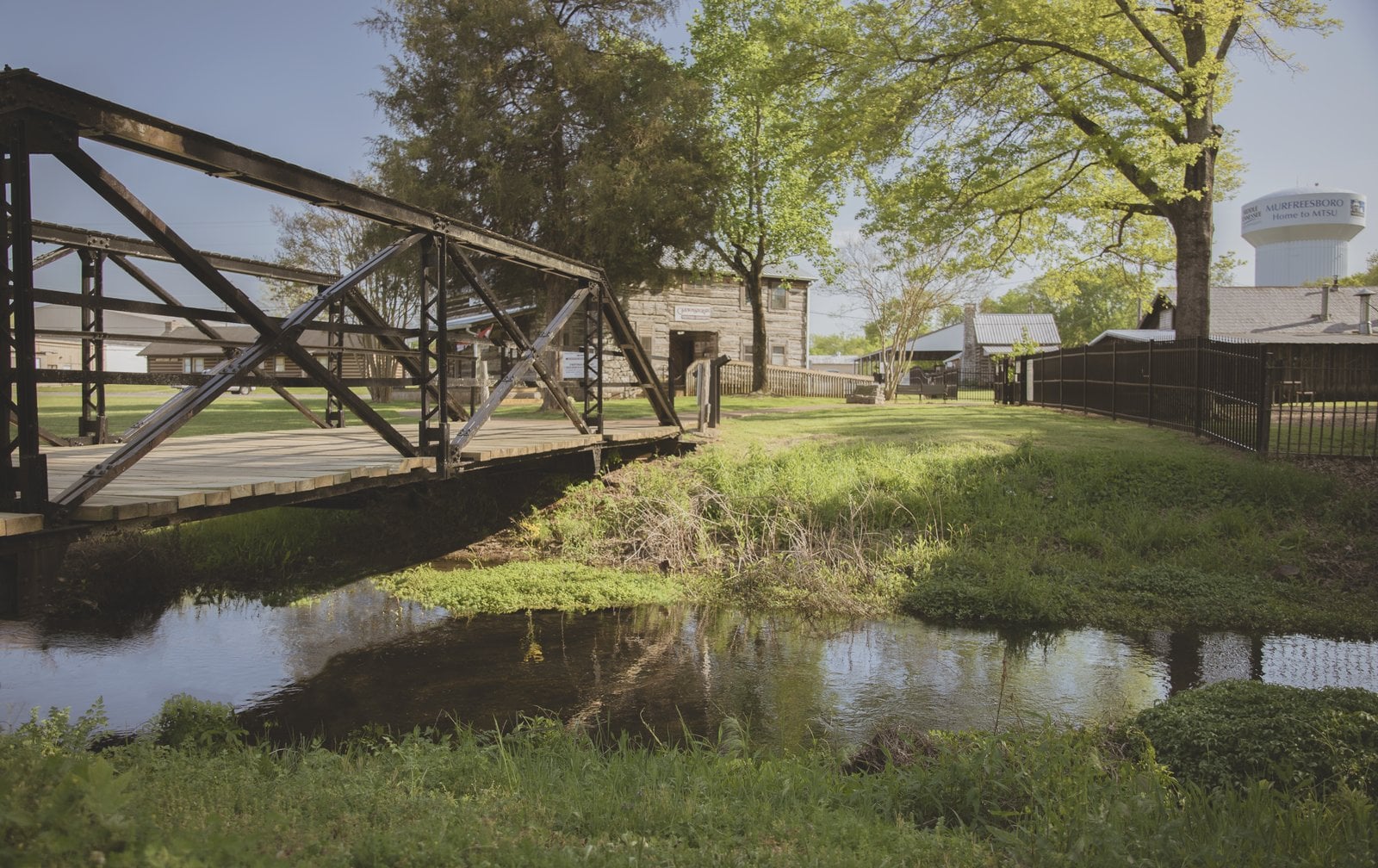Iron truss footbridge over a small creek, leading to a log building with a Cannonsburgh sign, with a Murfreesboro water tower behind the trees