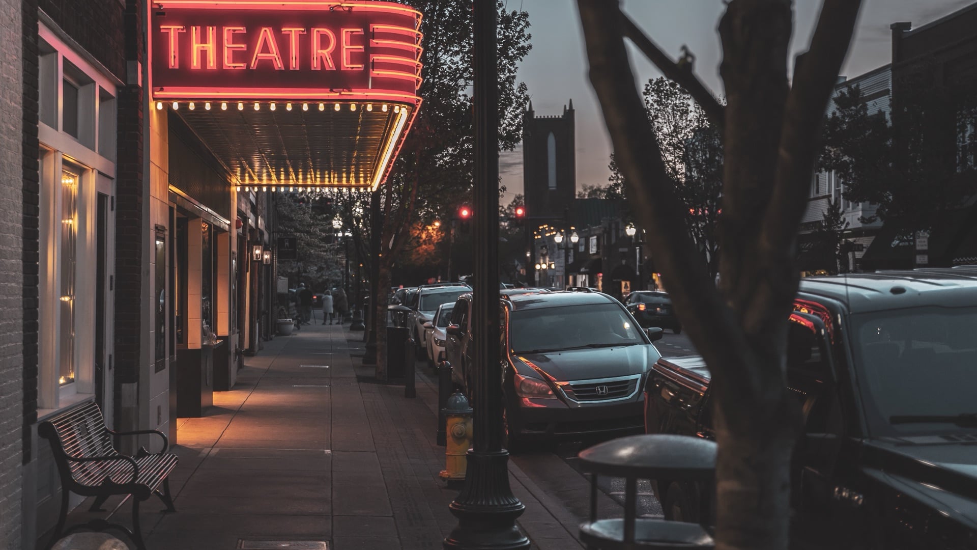 Lit Franklin Theatre marquee over the Main Street sidewalk at dusk, with a bench, parked cars and a stone tower down the street