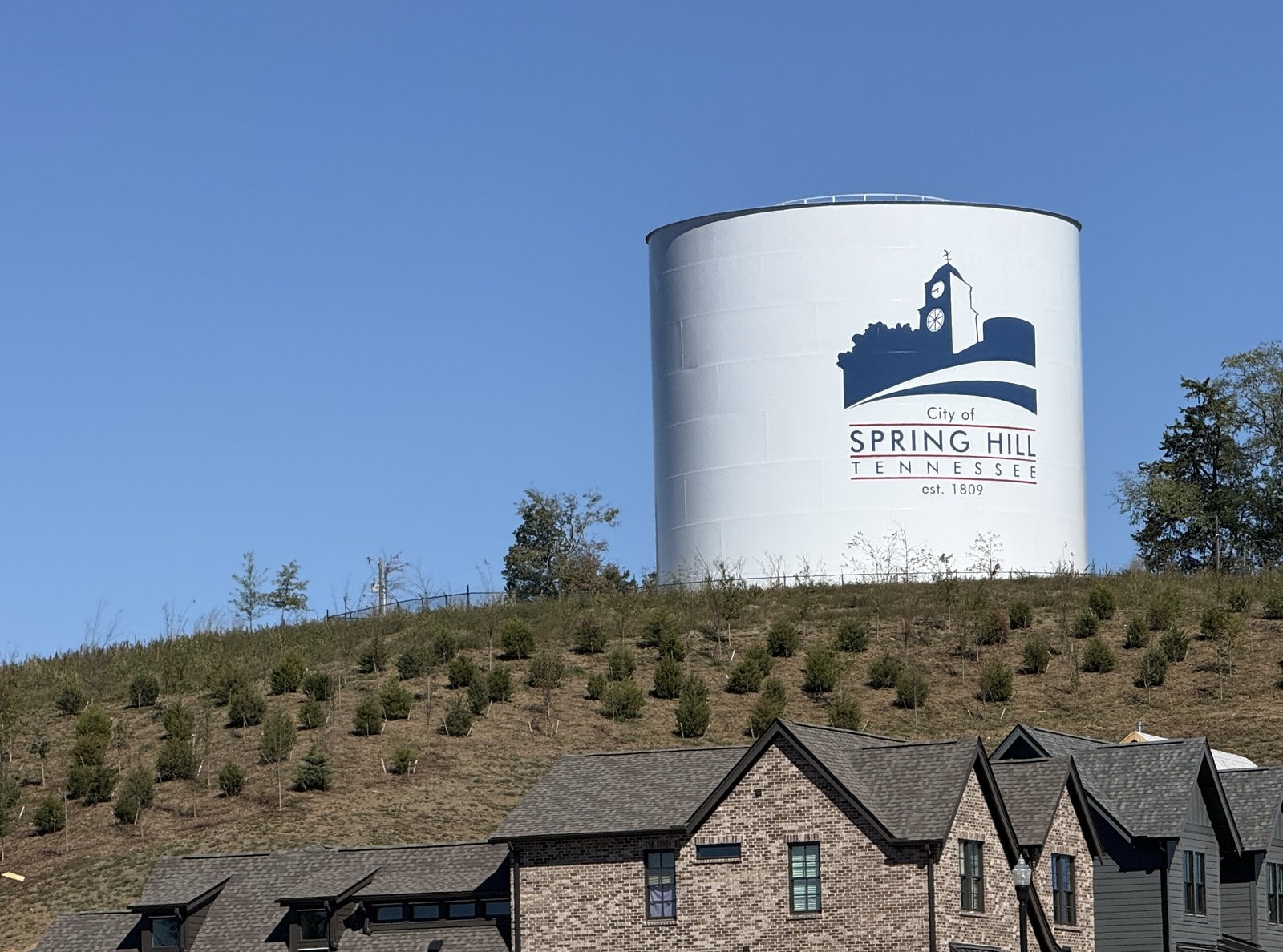 White City of Spring Hill water tank on a planted hillside above the rooflines of brick homes.