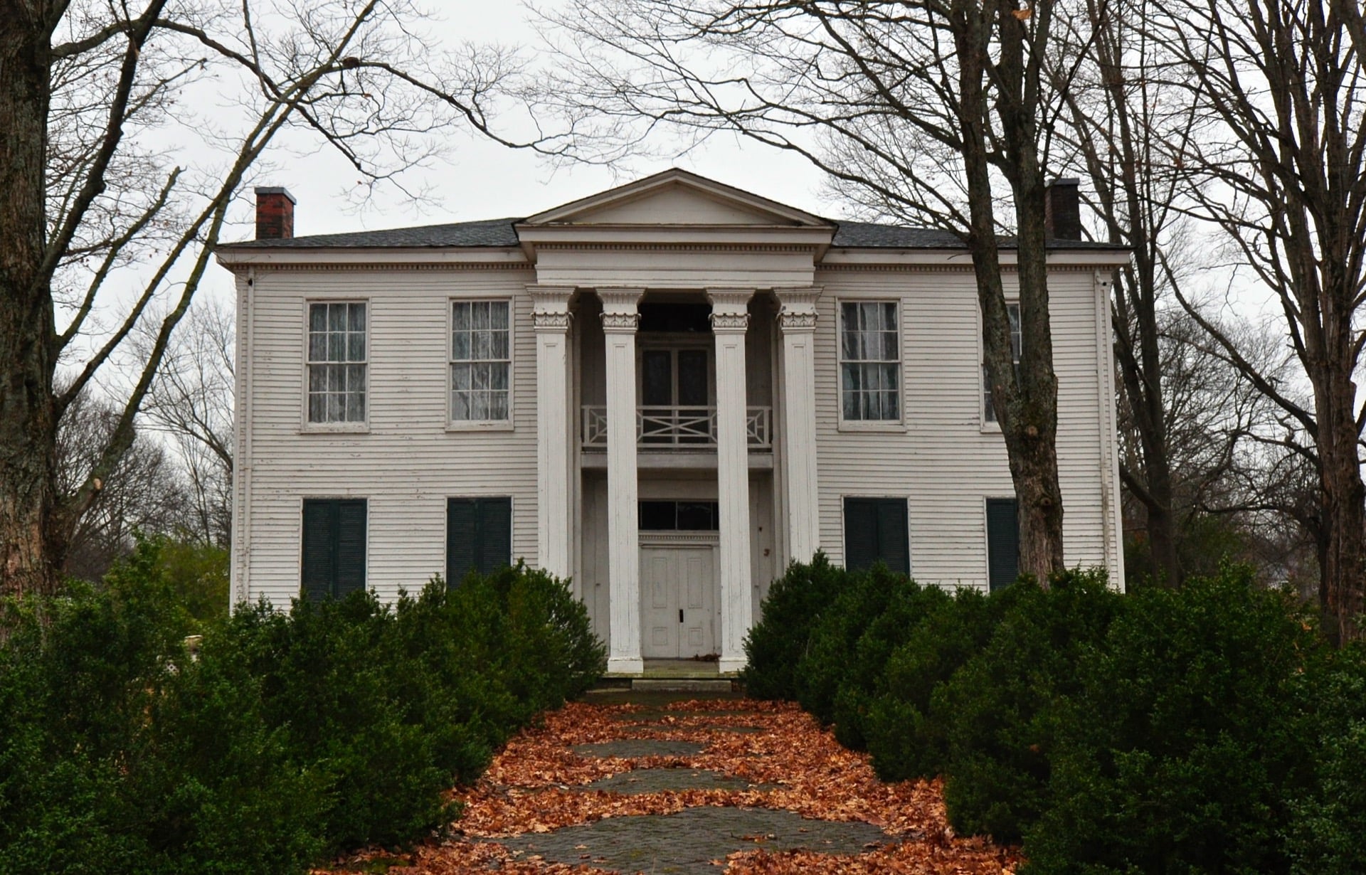 White Hall, a two-story white frame house with a columned portico and green shutters, behind boxwood hedges and a leaf-covered stone walk