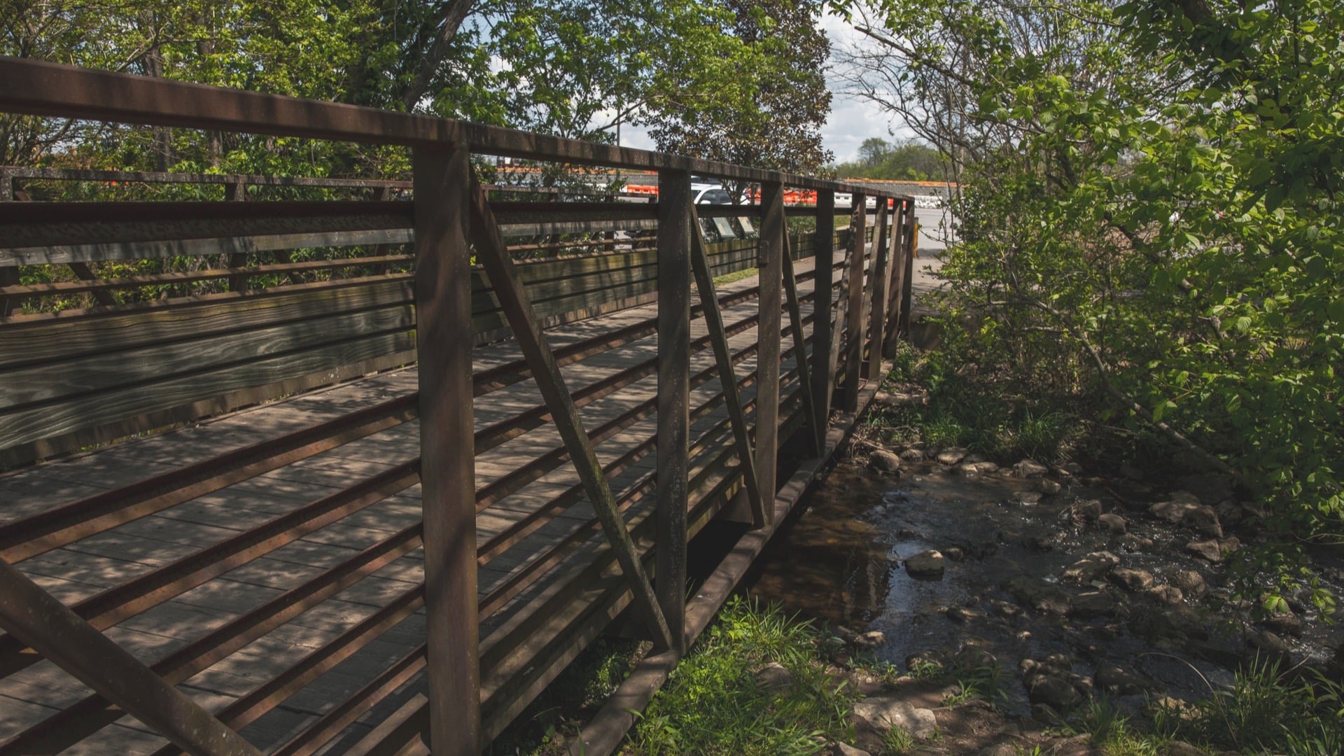 Wooden footbridge with steel railings over a shallow, rocky creek, with trees on both banks