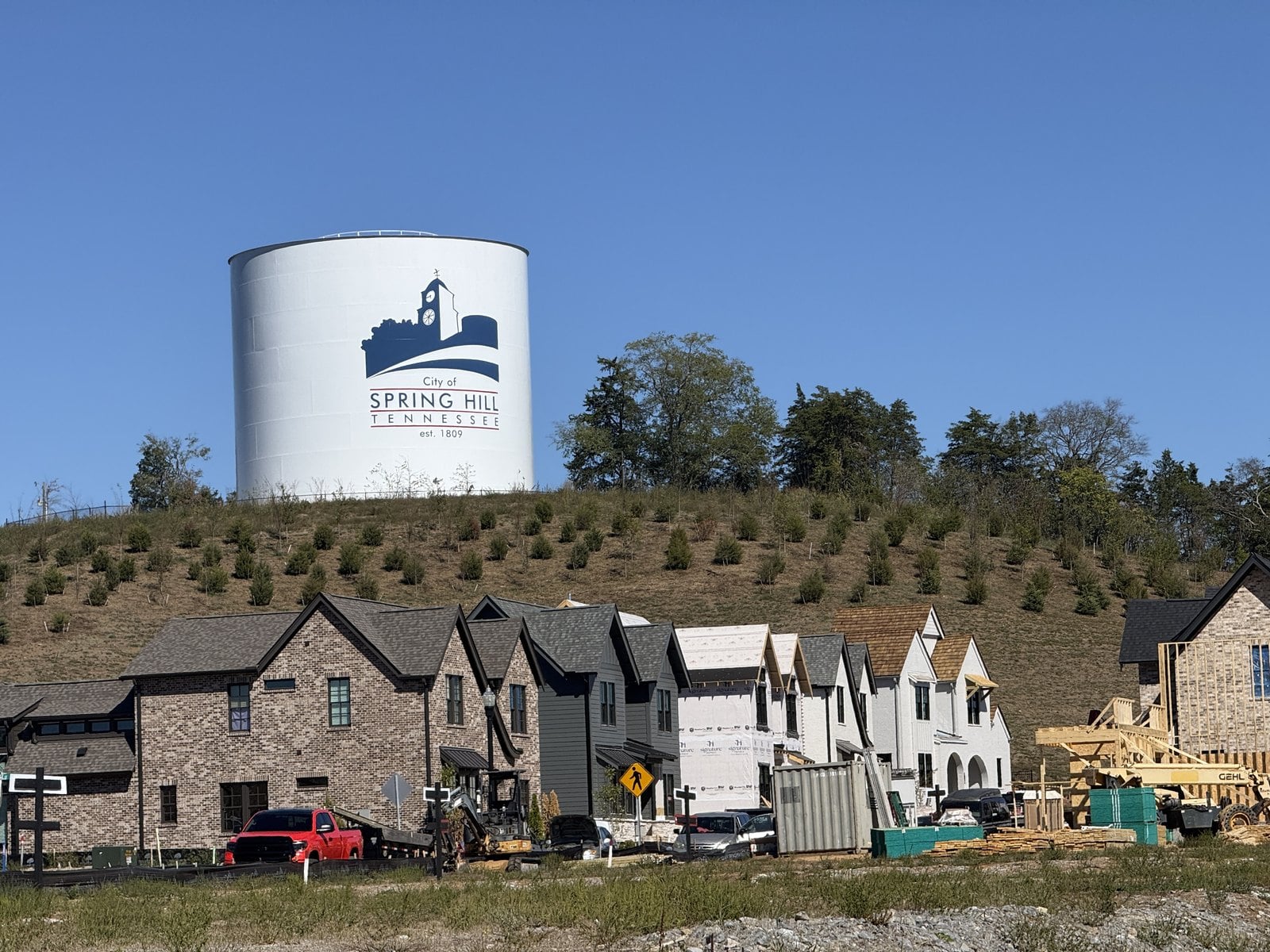 The white City of Spring Hill water tower on a hill above a row of new brick and white homes under construction in June Lake, Spring Hill, TN