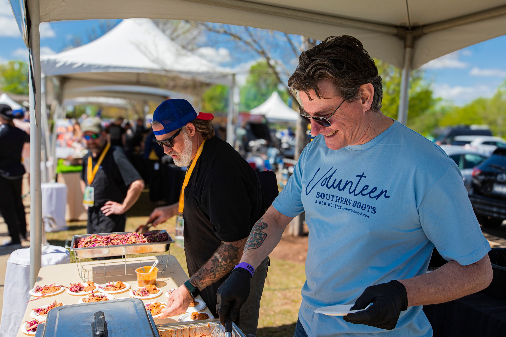 Two volunteers serve plates of food under a white tent at the Southern Roots Reunion event. One man in a blue “Volunteer – Southern Roots BBQ Reunion” shirt plates food from a tray while another prepares dishes in the background. Outdoor food festival setting.