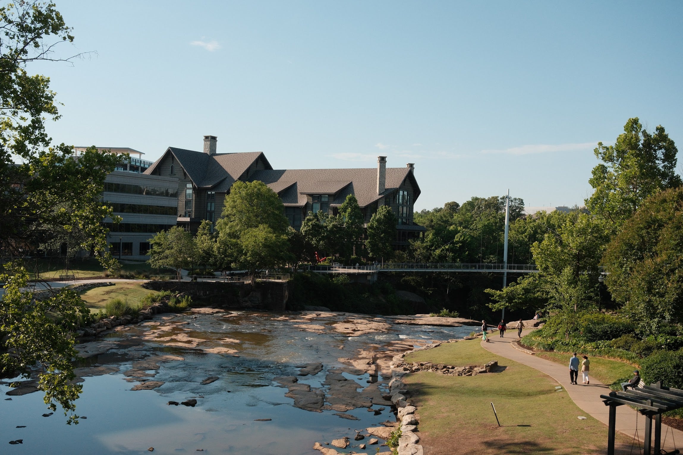 View of Falls Park on the Reedy in downtown Greenville, South Carolina, featuring the Liberty Bridge, Reedy River, walking trails, and visitors enjoying the park.