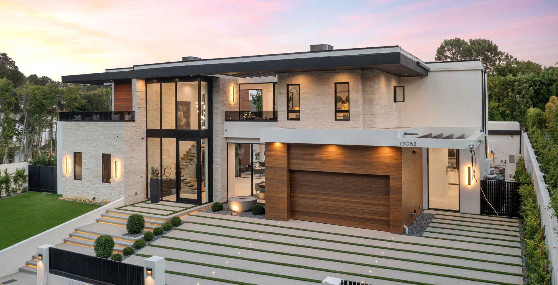 Modern luxury home exterior with large windows, wooden garage door, and well-manicured lawn at sunset.