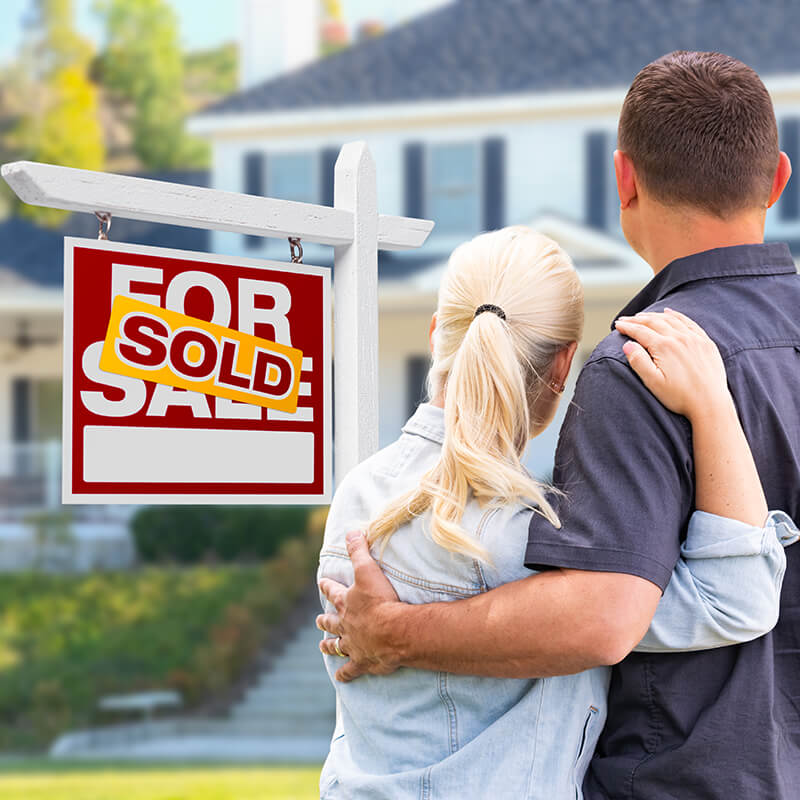Young Adult Couple Facing Front of Sold Real Estate Sign and House