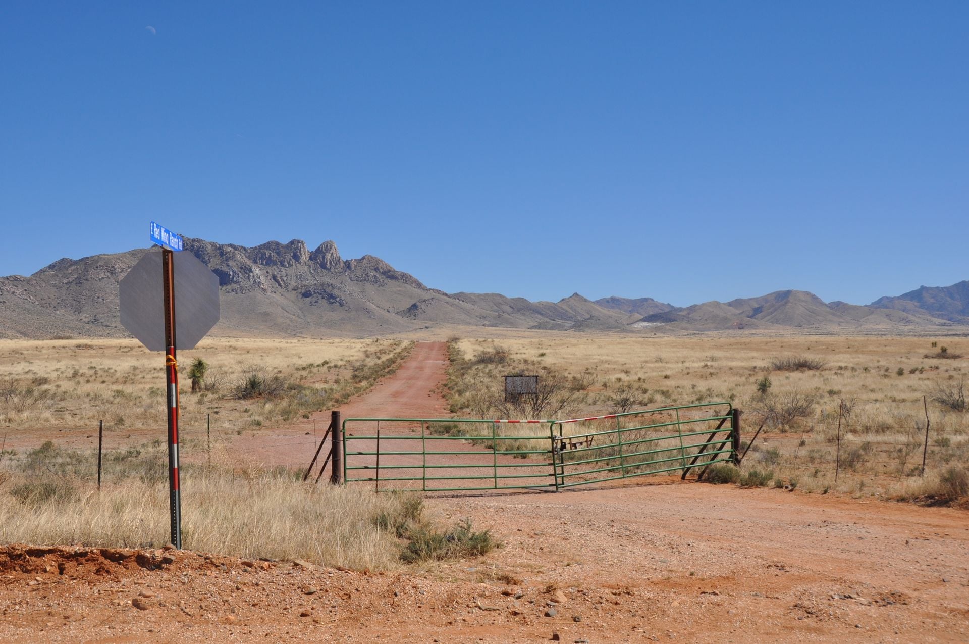 Green ranch gate across a red dirt road leading to open Texas land and mountains under a clear blue sky