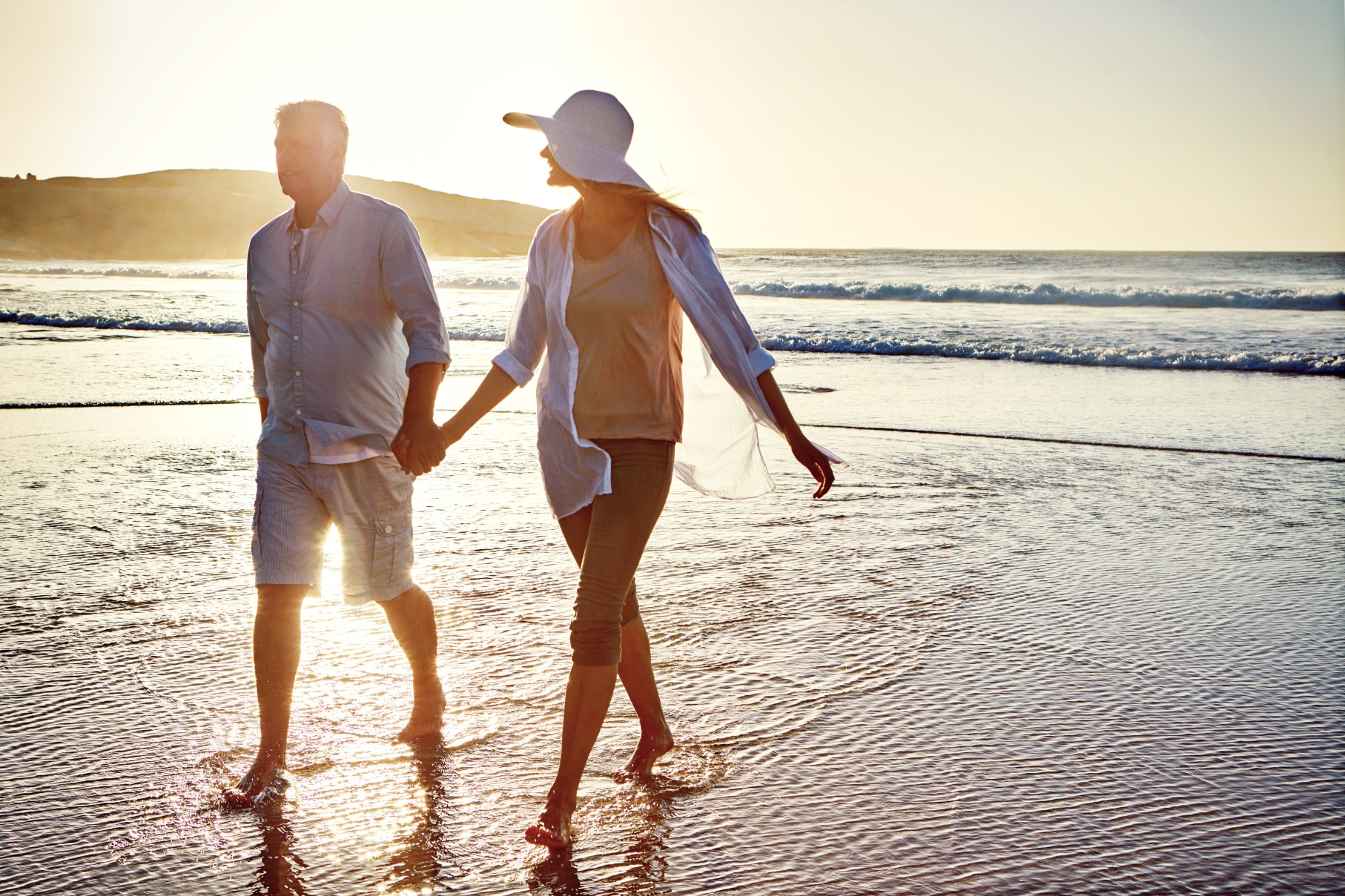Senior couple, holding hands and walking with beach sunset for bonding, love or support together in nature. Man, woman or lovers enjoying stroll on outdoor summer holiday by water, sea or ocean coast