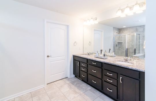This is an image of a bathroom. It shows a double vanity with a granite countertop, a large mirror, two sinks, a shower stall, and a white door