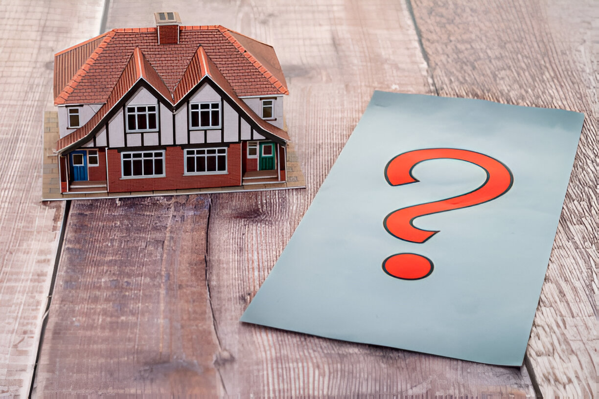 The image contains a miniature model house and a piece of paper with a red question mark on it.