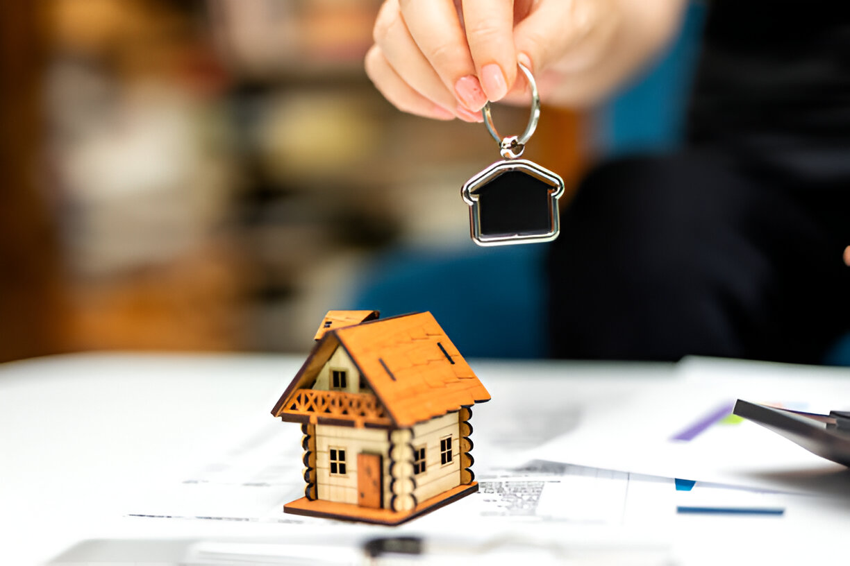 The image shows a hand holding a house-shaped keychain above a small wooden model house