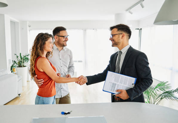 image shows a real estate agent shaking hands with a couple in what appears to be an empty apartment