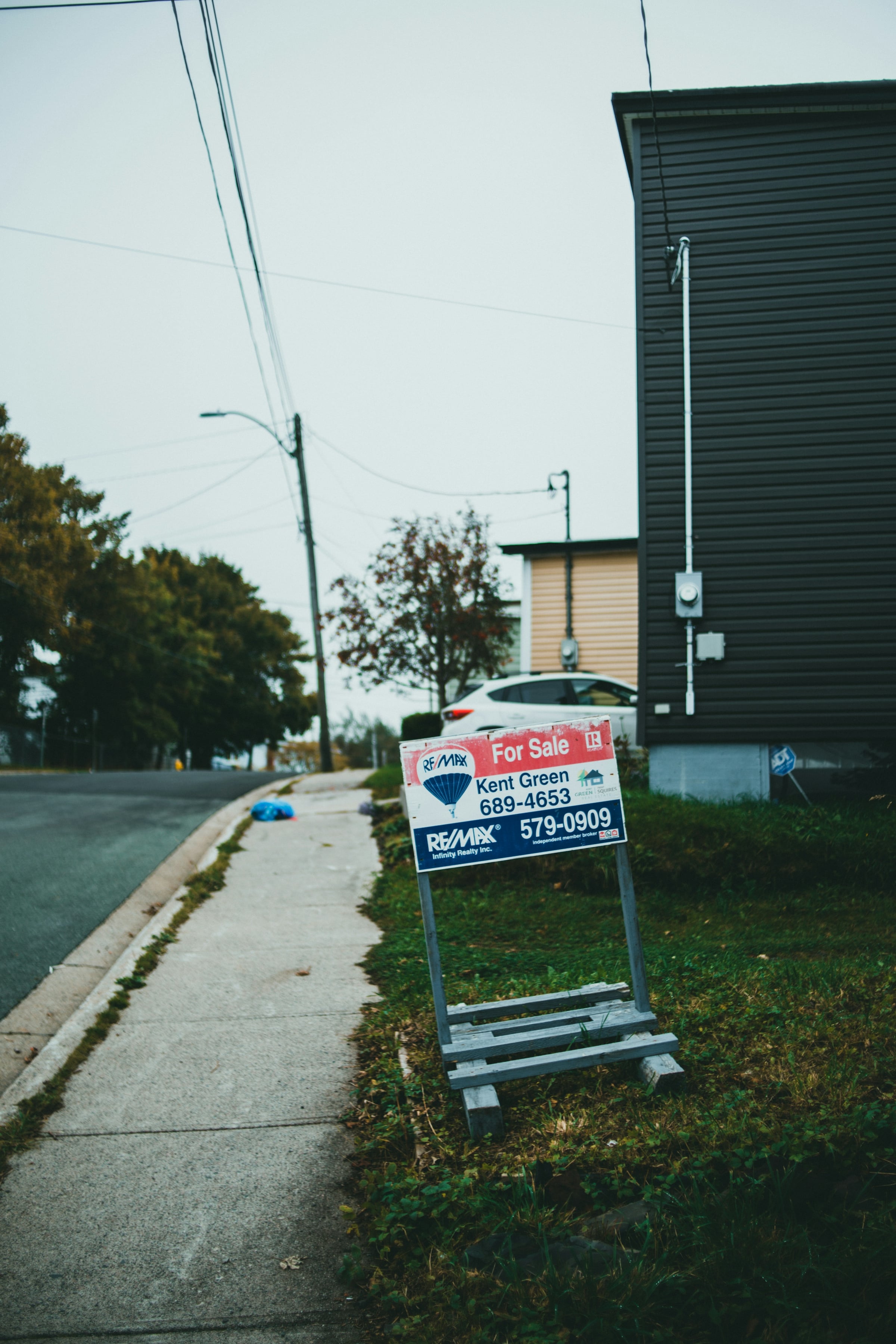 Real estate for-sale yard sign staked in a grass strip beside a sidewalk in front of a house