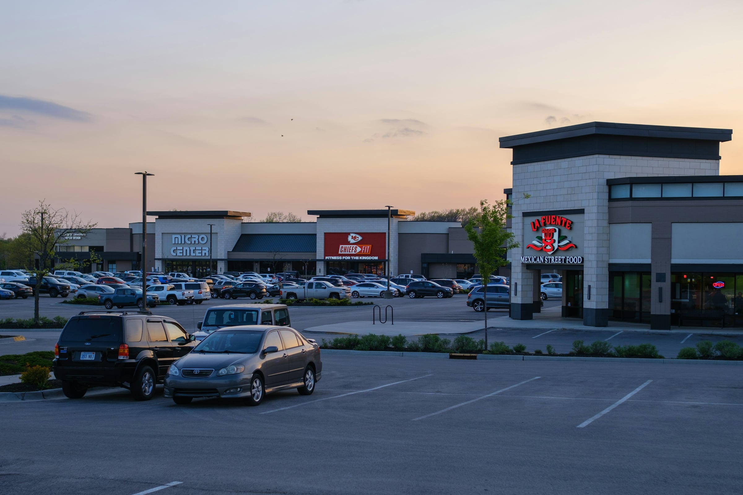 Suburban retail strip center at dusk with lit storefront signage and a striped parking lot