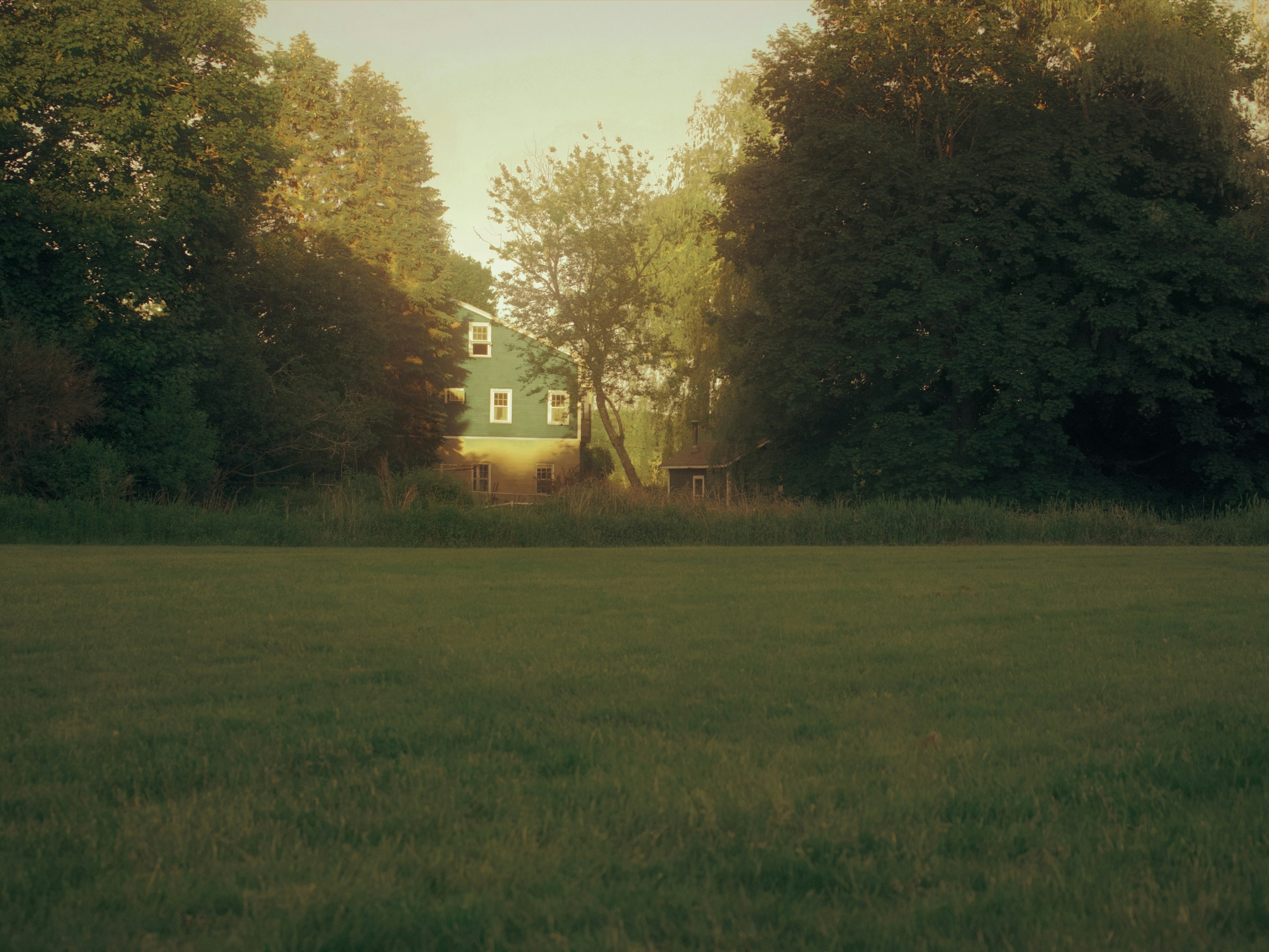 Open grassy undeveloped parcel flanked by deciduous woods with a single house at the far treeline