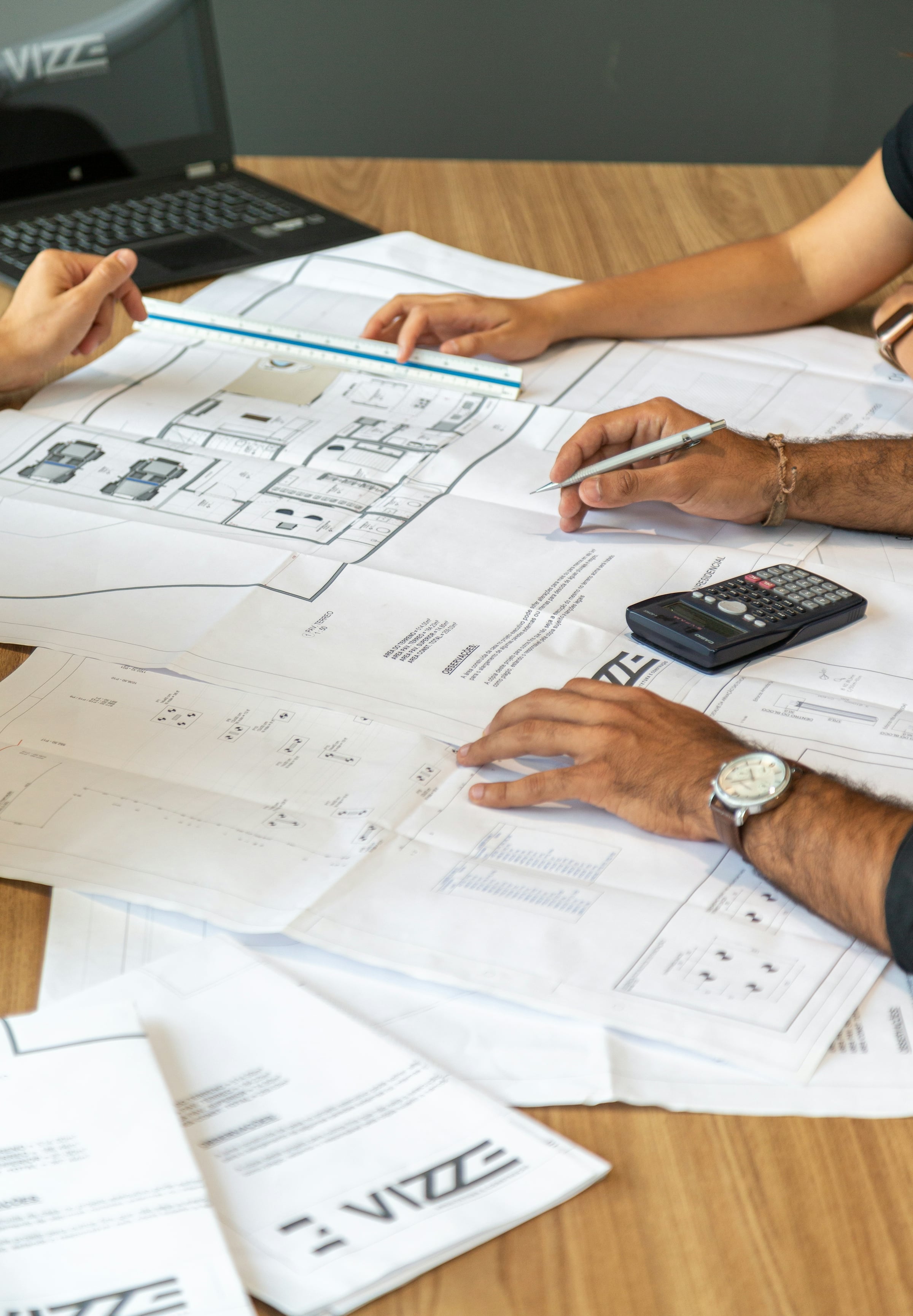 Two people marking up architectural floor plans spread across a wood table, with a calculator beside them