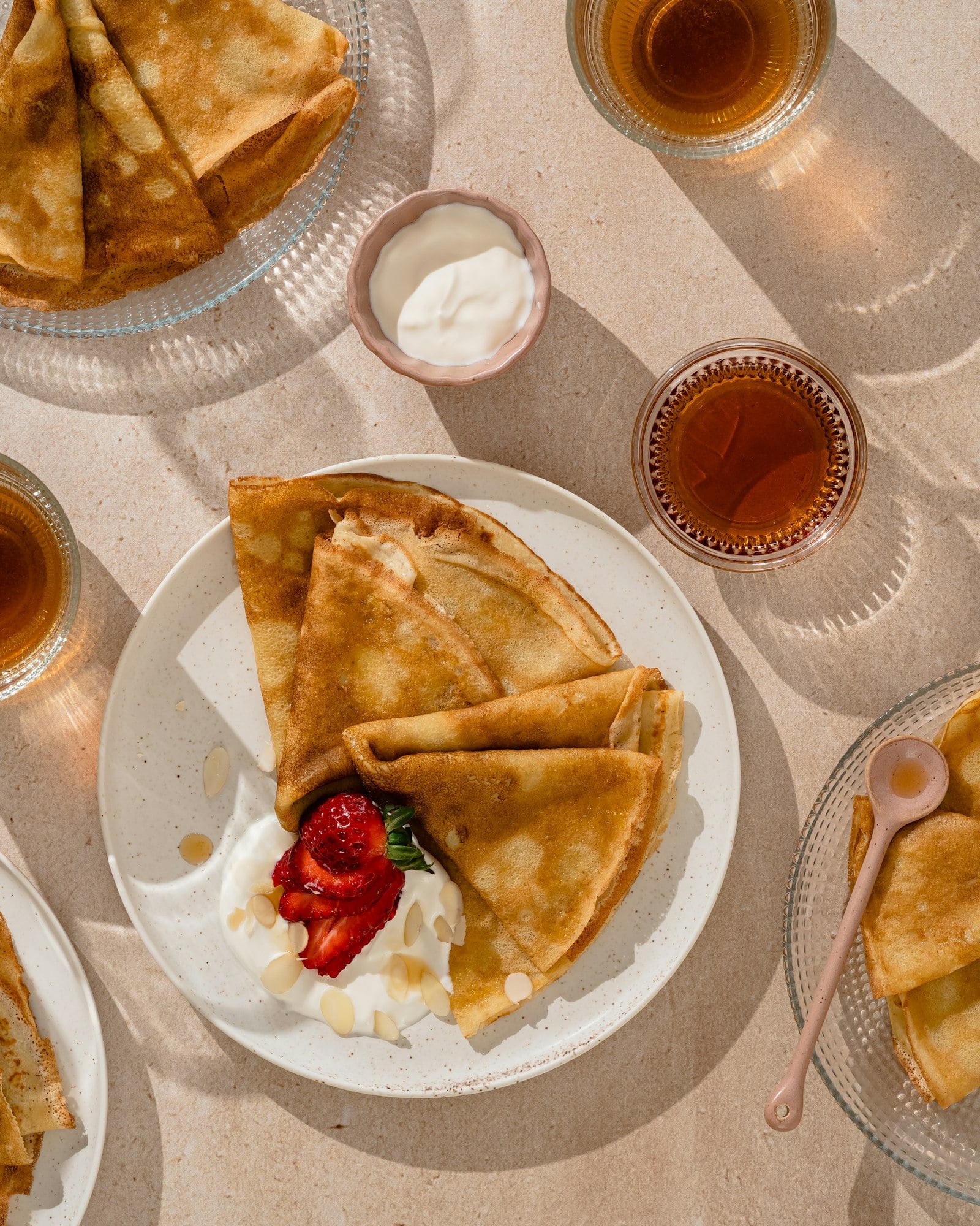Overhead view of folded crepes with strawberries, yogurt and small glasses of syrup on a sunlit table, set for a brunch spread.