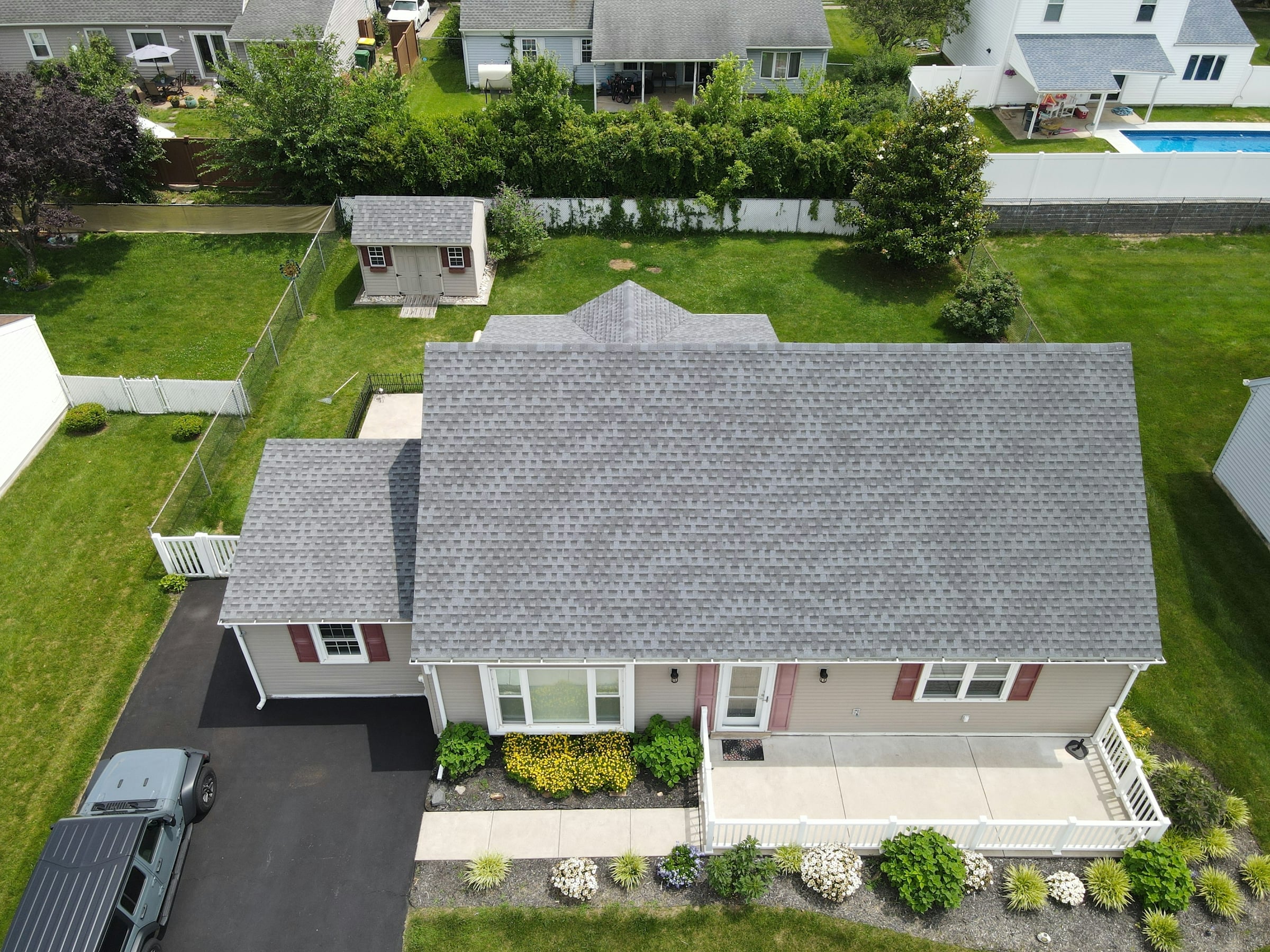 An aerial view of a single-family house with a grey asphalt shingle roof, a paved driveway and a mown back lawn.