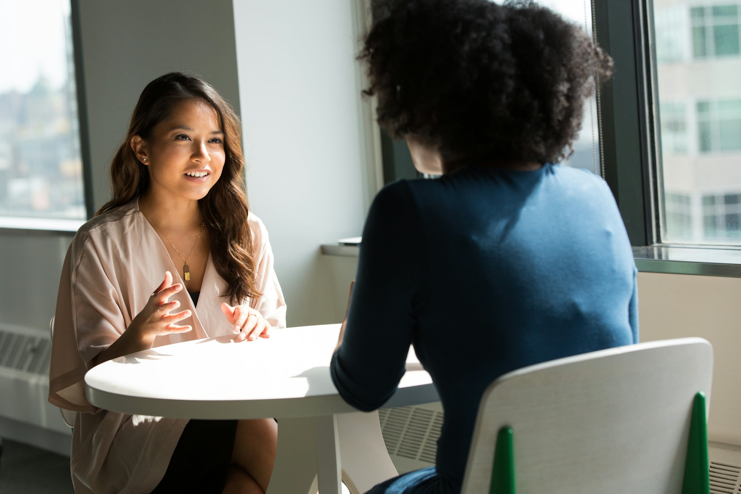 A woman seated at a small table talking with someone across a laptop