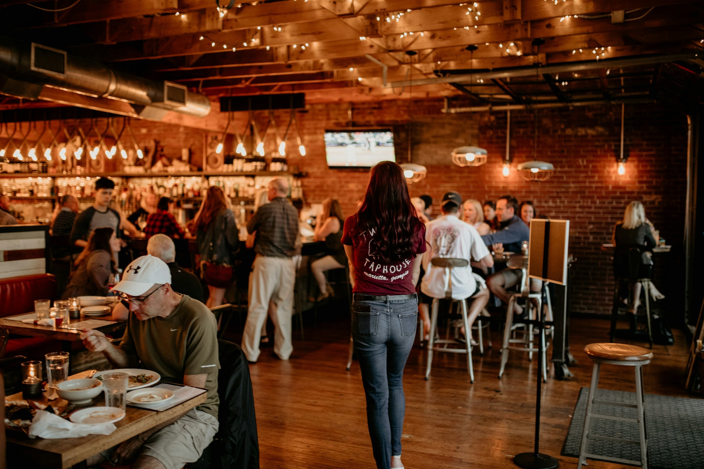 A busy brewery taproom with exposed brick walls, pendant lights and customers standing and sitting at high tables.