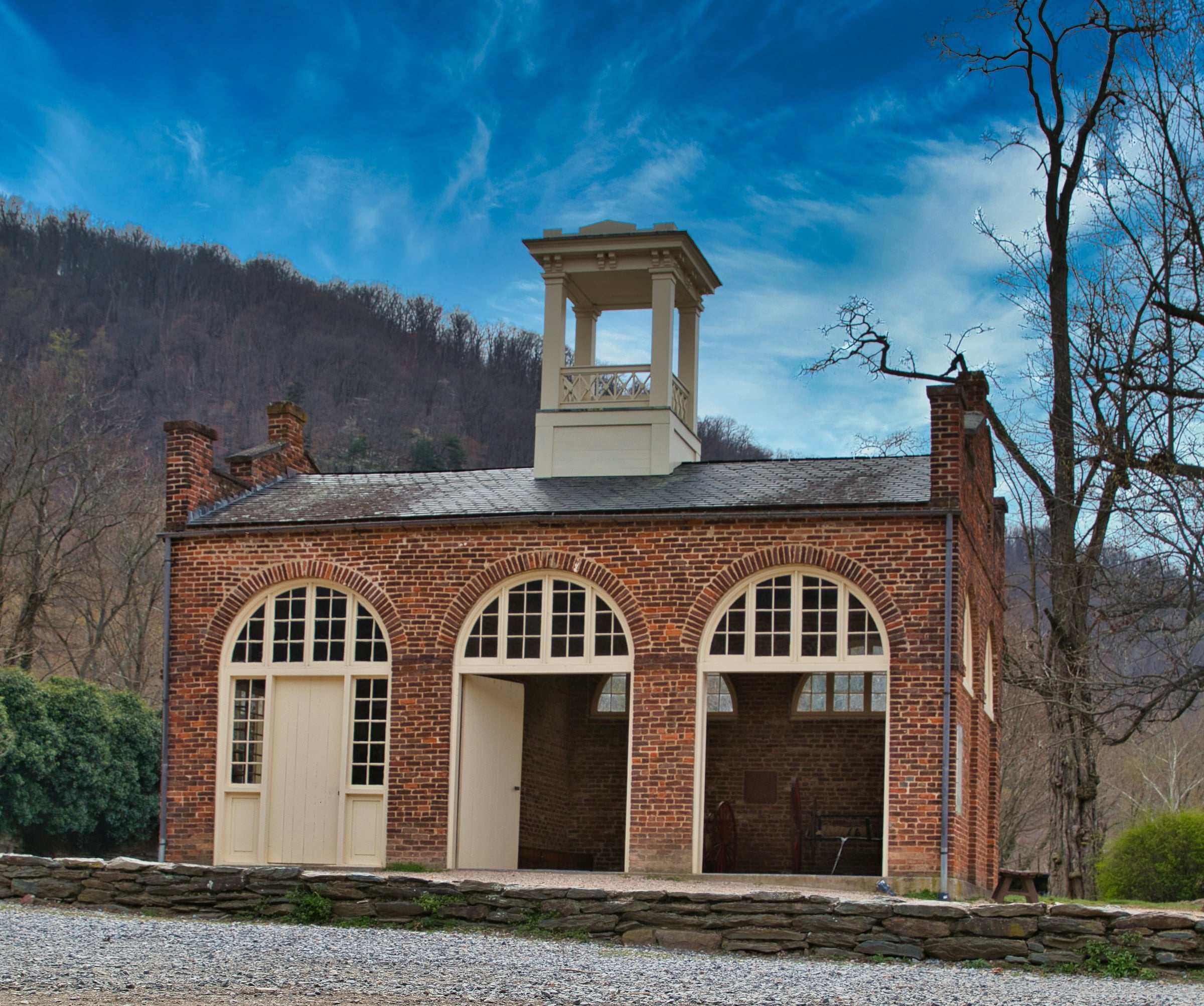An old brick building with three arched bay doors and a rooftop cupola, set against a wooded hillside.