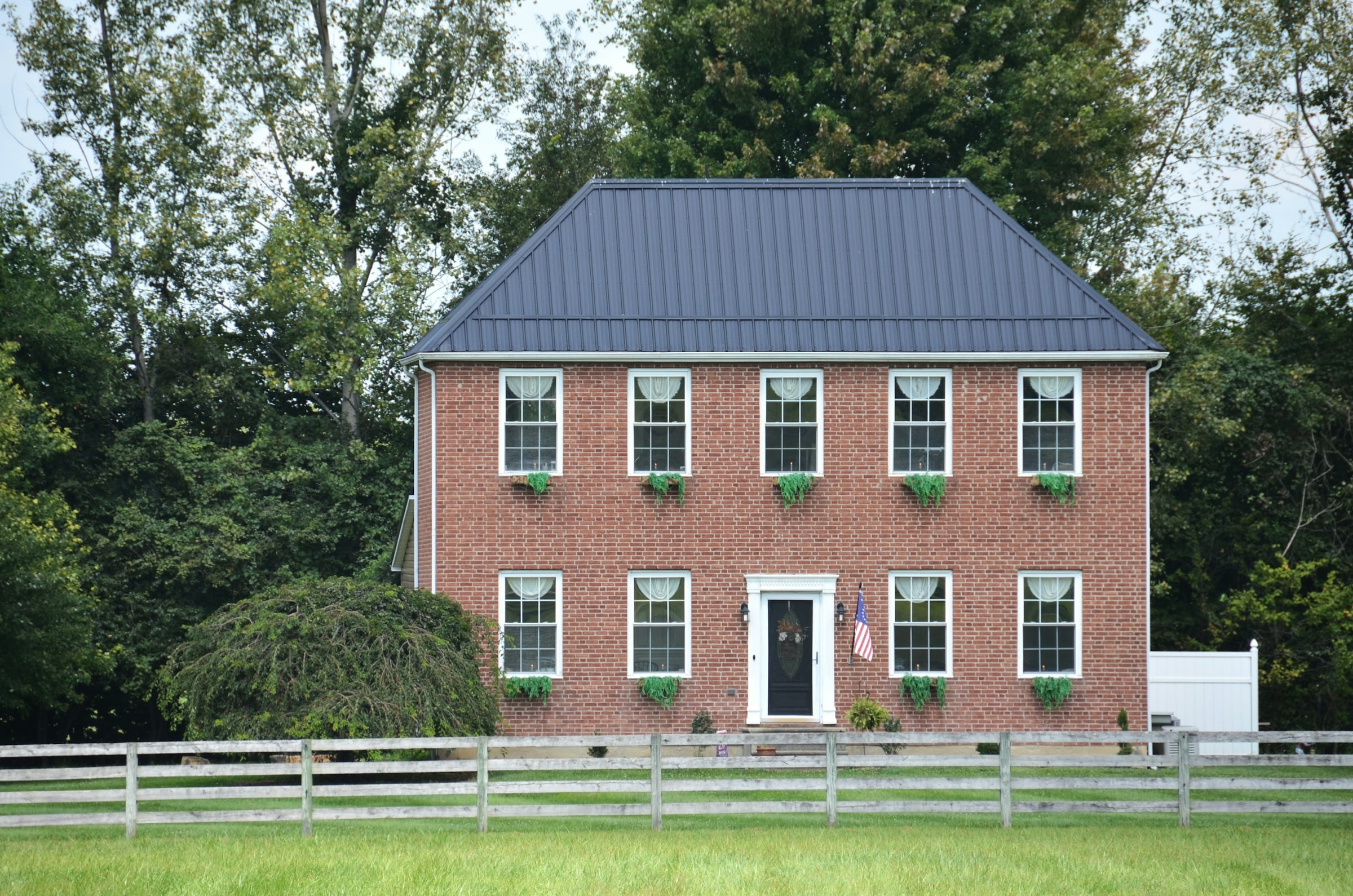 A two-story red brick colonial house with black shutters and a dark metal roof, seen across a mown lawn behind a white rail fence.