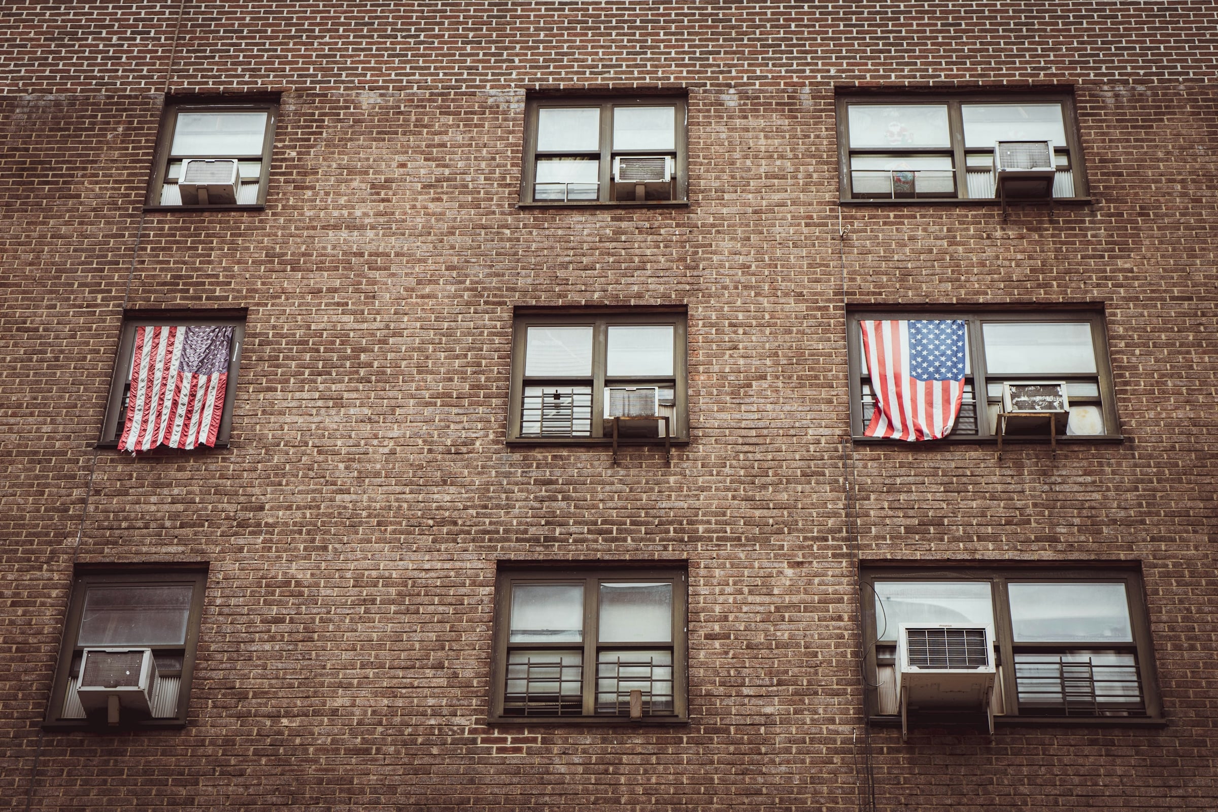 The brick facade of a low-rise condominium building, with window air-conditioning units and United States flags hanging outside several units.