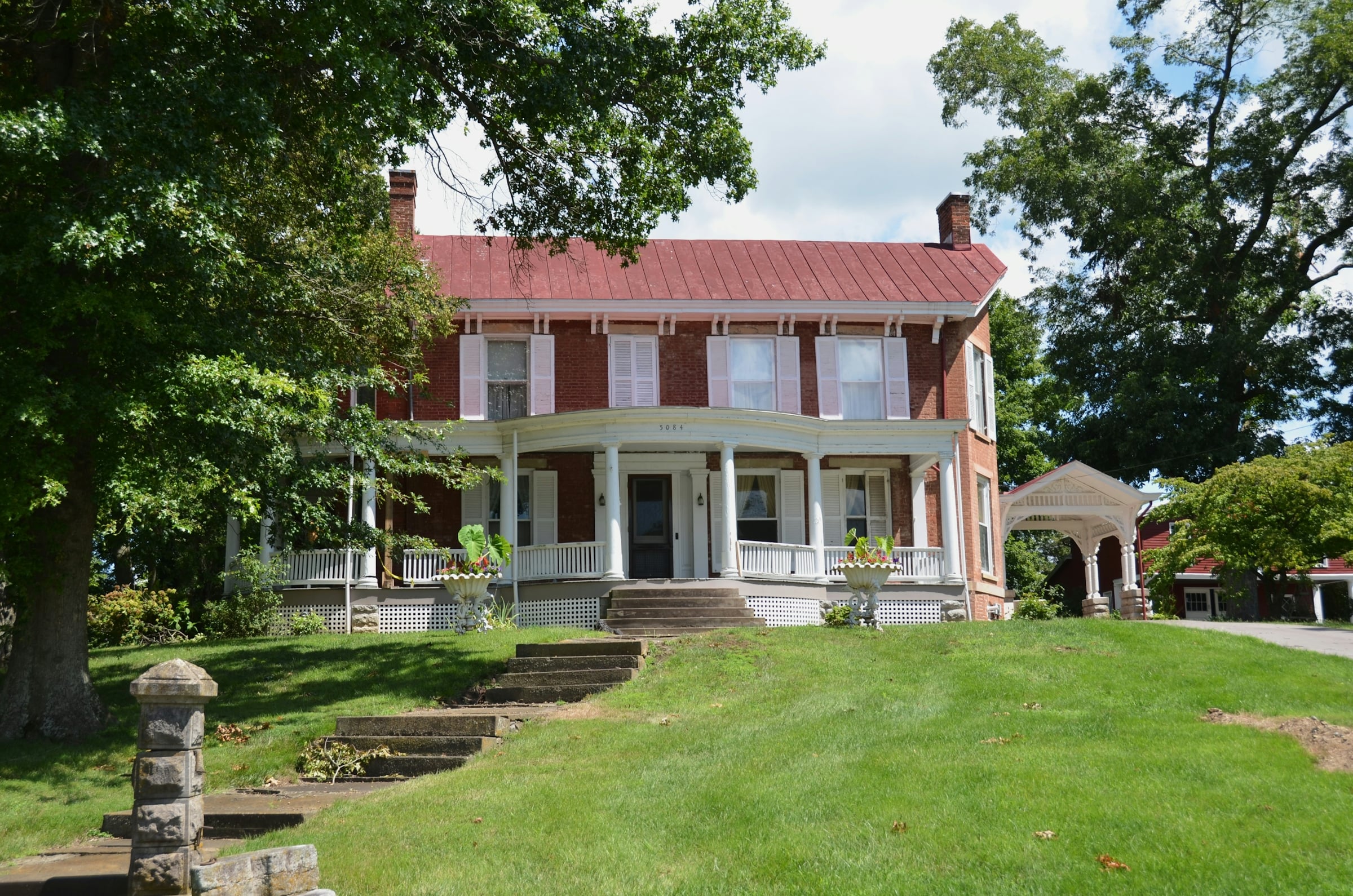A large two-storey brick house with a metal roof and white columns set back behind a wide mown lawn and mature trees