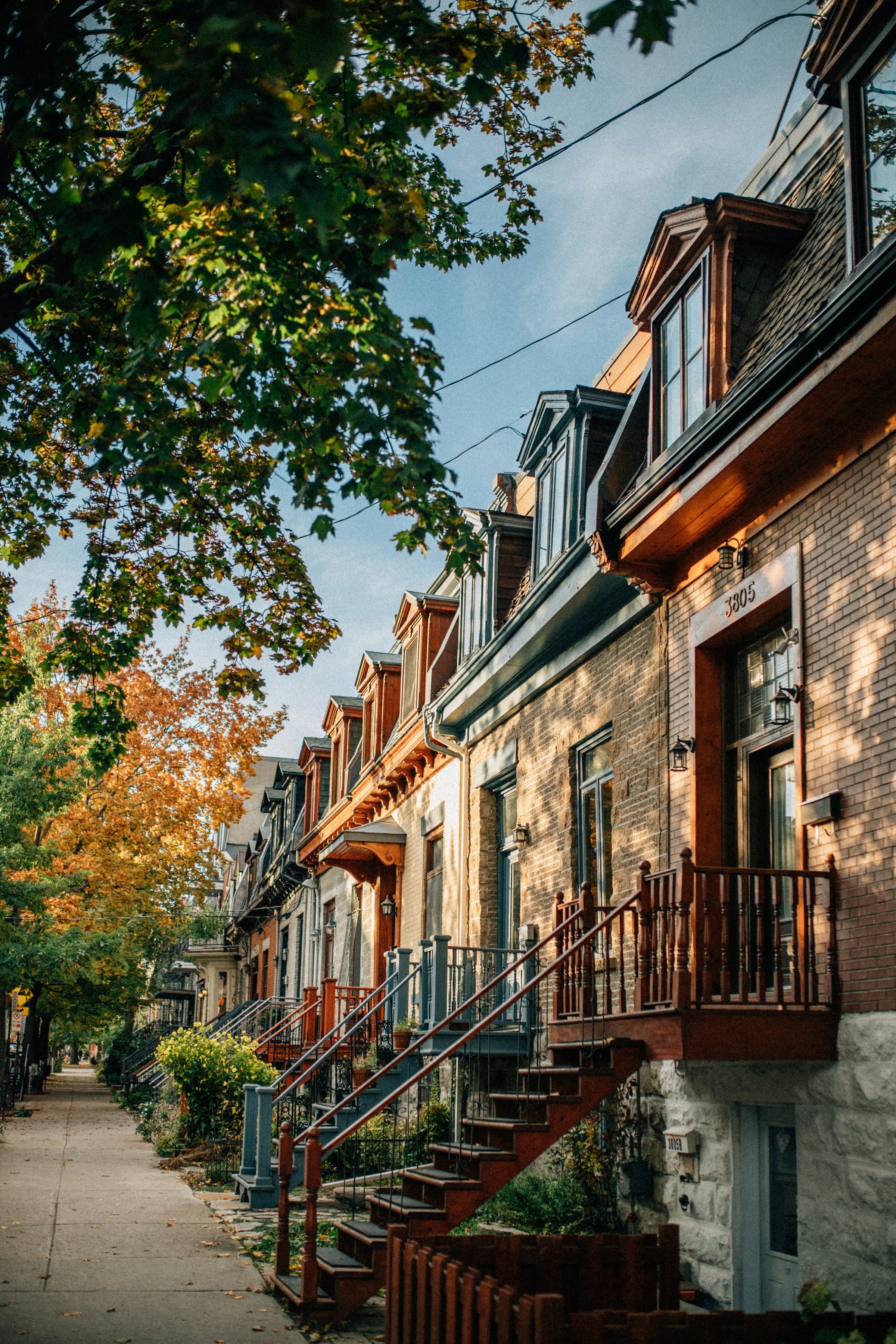 A row of attached brick townhouses with green window awnings under a turning autumn tree.