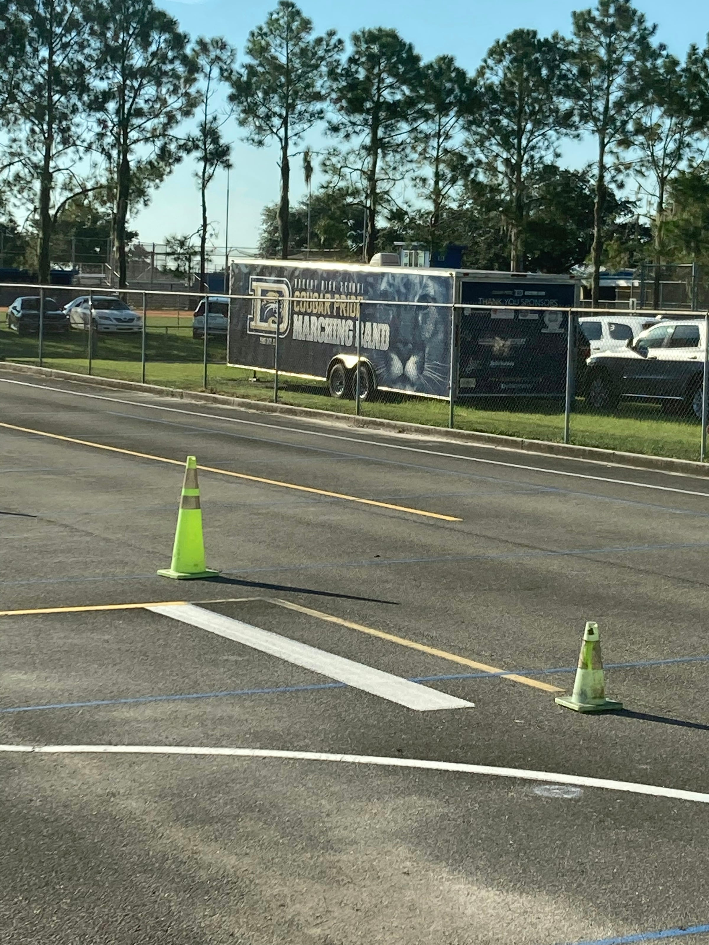 A cleared paved lot behind a chain-link fence, with traffic cones on the roadway and mature trees along the back boundary.