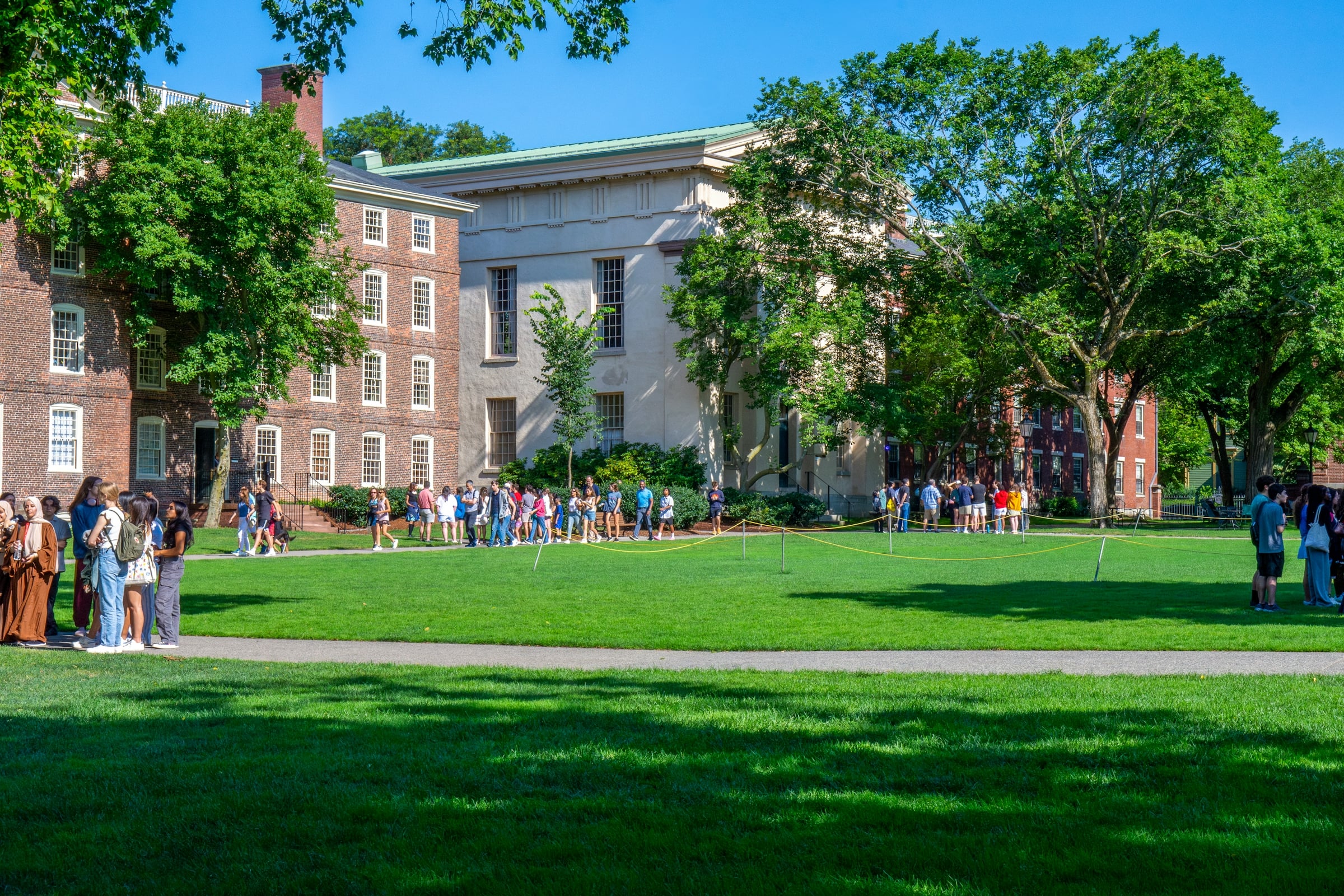 A brick college building beside a wide green lawn where a crowd of people has gathered.