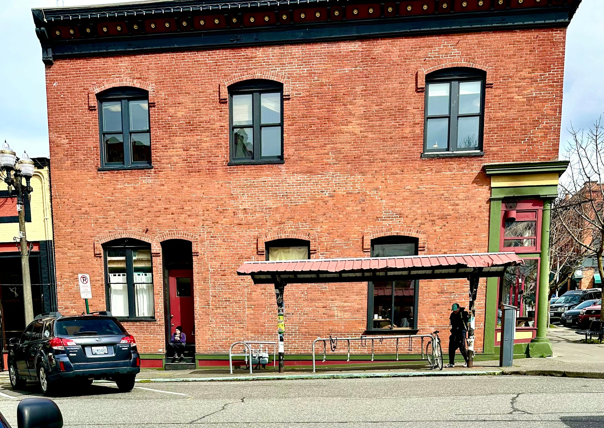 A two-story red brick commercial building with a metal awning over the sidewalk entrance, a bike rack out front and street parking.