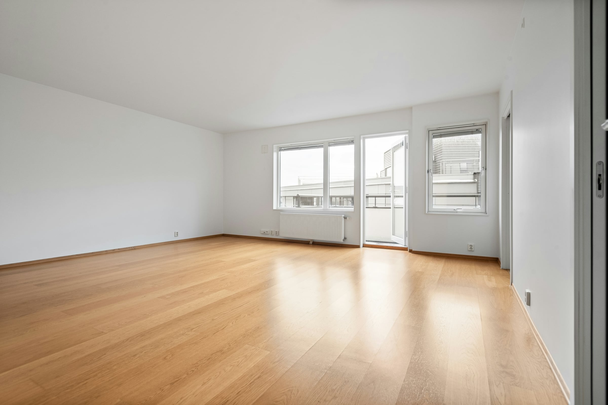 An empty room in a vacated house, with a pale hardwood floor, white walls and daylight coming through a bank of tall windows.