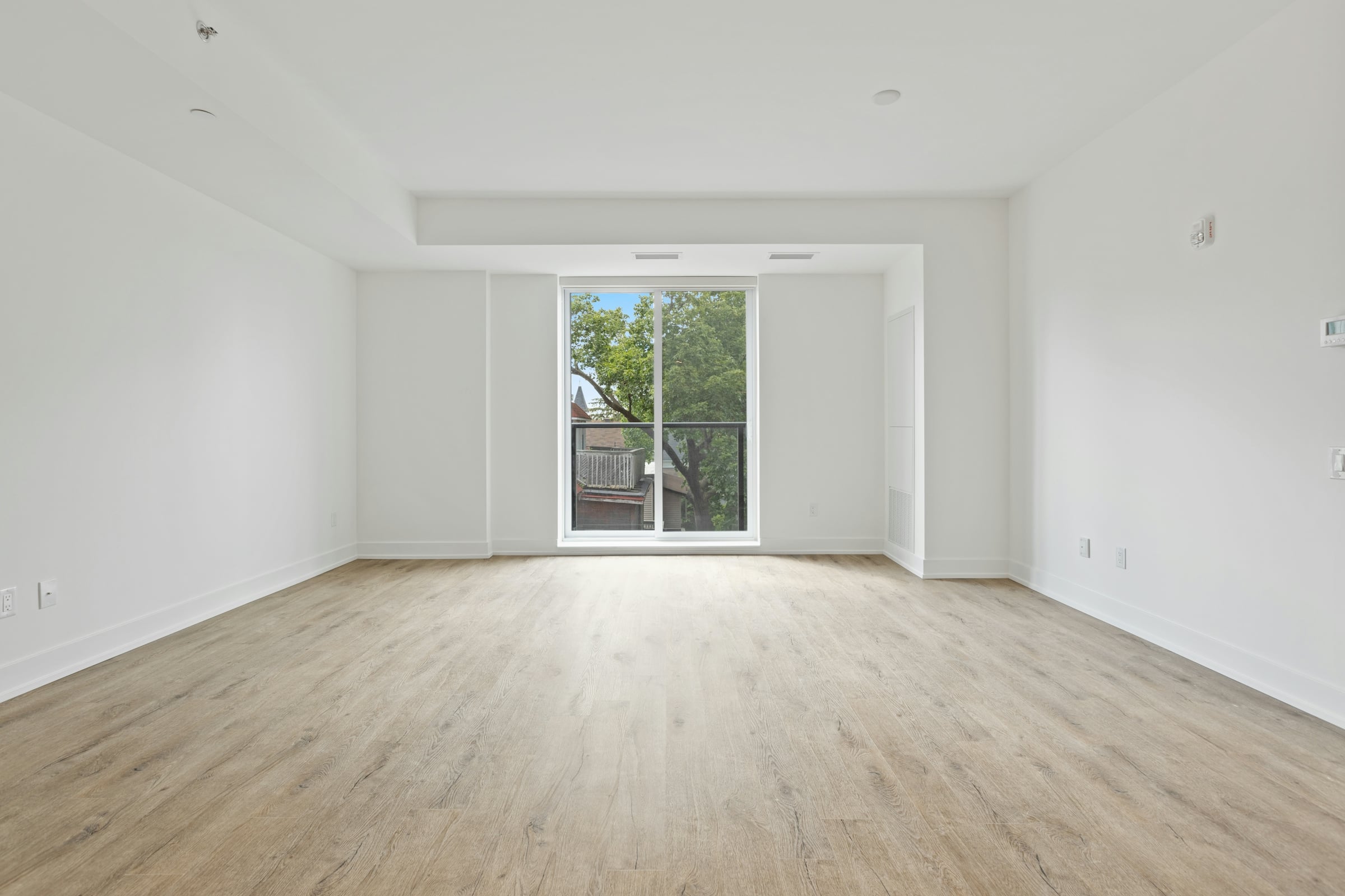 An empty room with pale wood flooring, bare white walls and a sliding patio door onto a deck