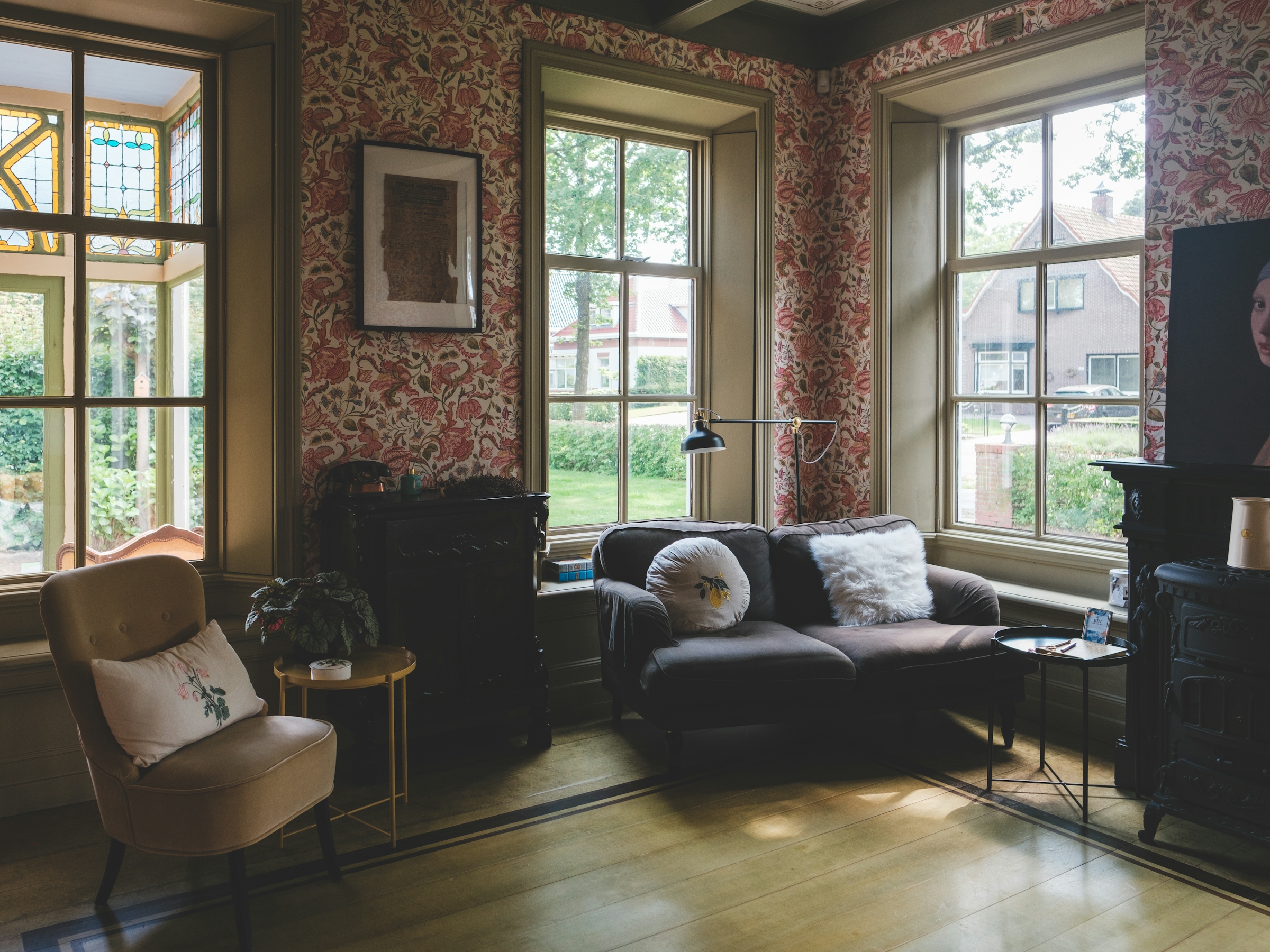 A furnished older living room with patterned wallpaper, tall windows, an armchair and a sofa lit by daylight.