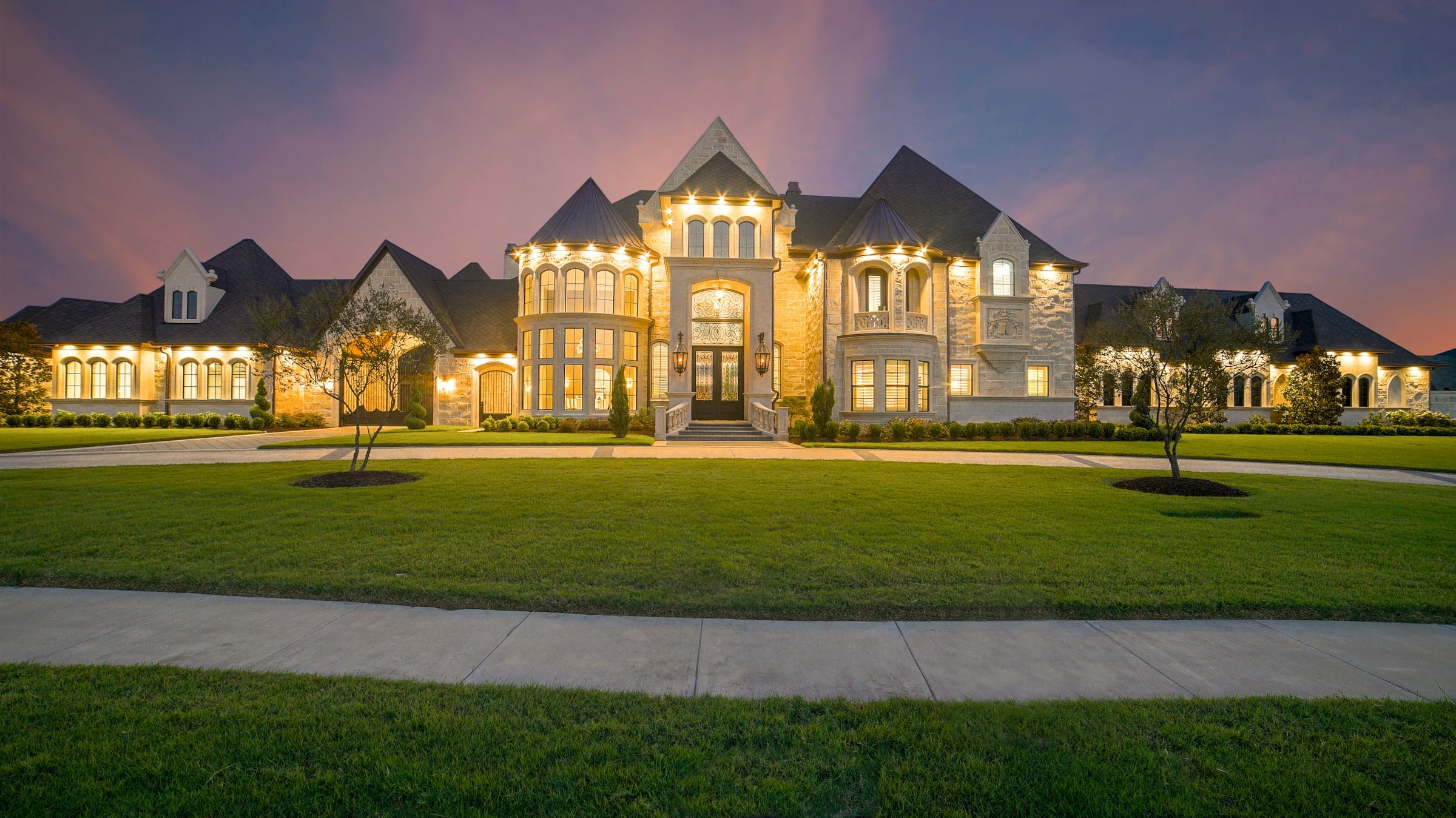 Large multi-gabled house lit from inside at dusk behind a wide manicured front lawn