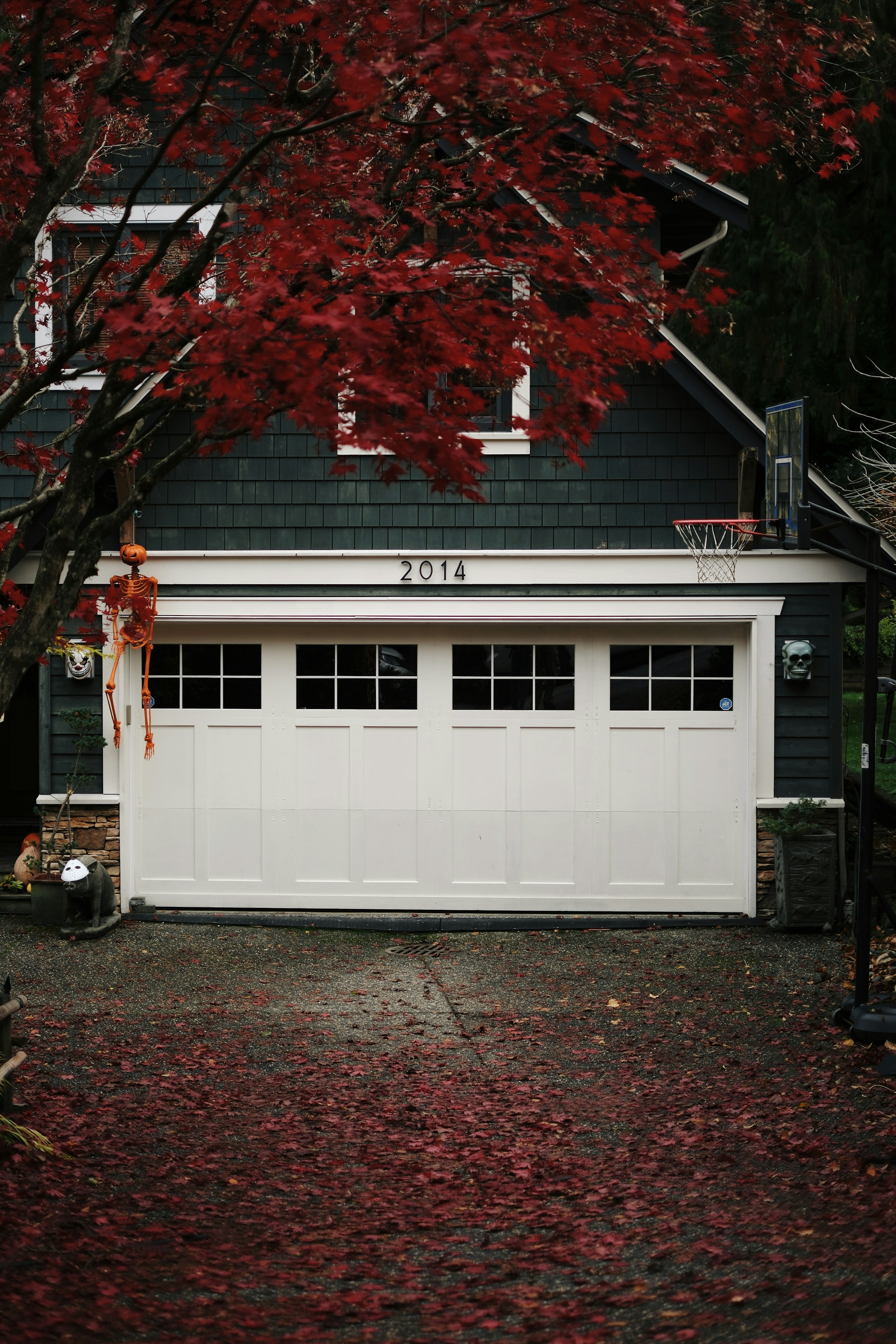 A white double garage door on a house with dark green siding, with fallen red maple leaves covering the driveway.